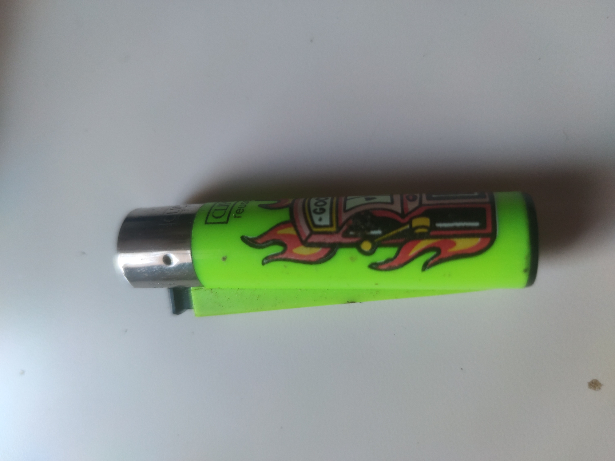 A lime green clipper lighter is positioned horizontally on a white surface. The lighter has a chrome top and is decorated with a cartoon graphic featuring flames and the word "GOON" in a stylized font. The lighter has the standard clipper design, including the removable flint housing. The surrounding surface is clean with a few small, dark specks. The city and specific location are Unknown.