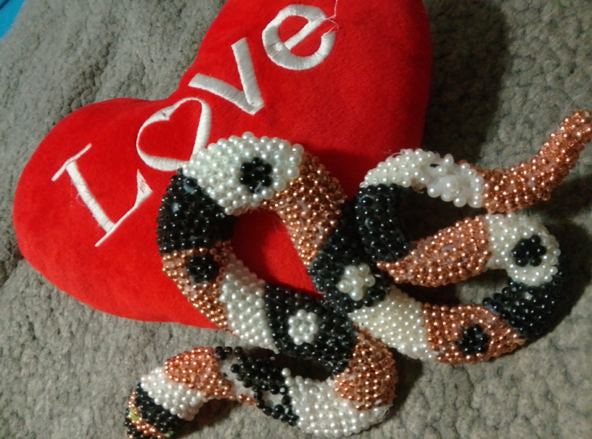 A red, heart-shaped pillow with the word "LOVE" embroidered in white is placed on a textured grey surface. A beaded object, made of white, black, and copper-colored beads, is intertwined with the pillow. The beaded object is shaped like a pretzel or a knot, with floral patterns created by the beads. The lighting suggests a close-up indoor shot, possibly taken during the daytime. There are no people or animals visible in the image. The specific location in Wedel, Germany is not ascertainable from the image content.