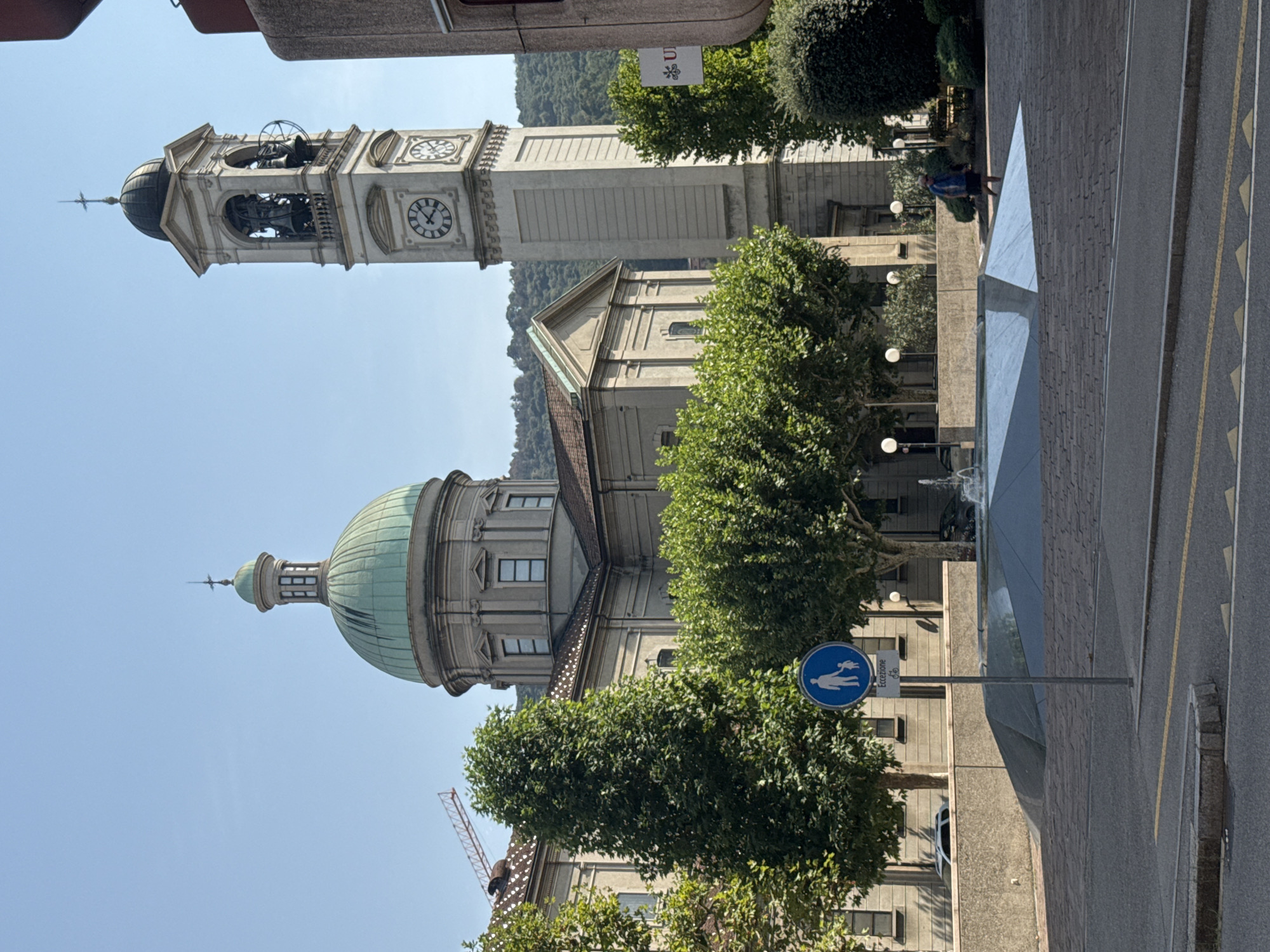 The image shows the center of Chiasso, Switzerland. A large dome and bell tower dominate the skyline. The dome is green, and the bell tower has a clock face and bells. In front of the buildings, there is a fountain and a sign with a pedestrian symbol. A person is walking in the distance. The area is paved with cobblestones. Trees and bushes surround the buildings. The sky is clear and blue. A construction crane is visible in the background, suggesting ongoing construction or renovation.