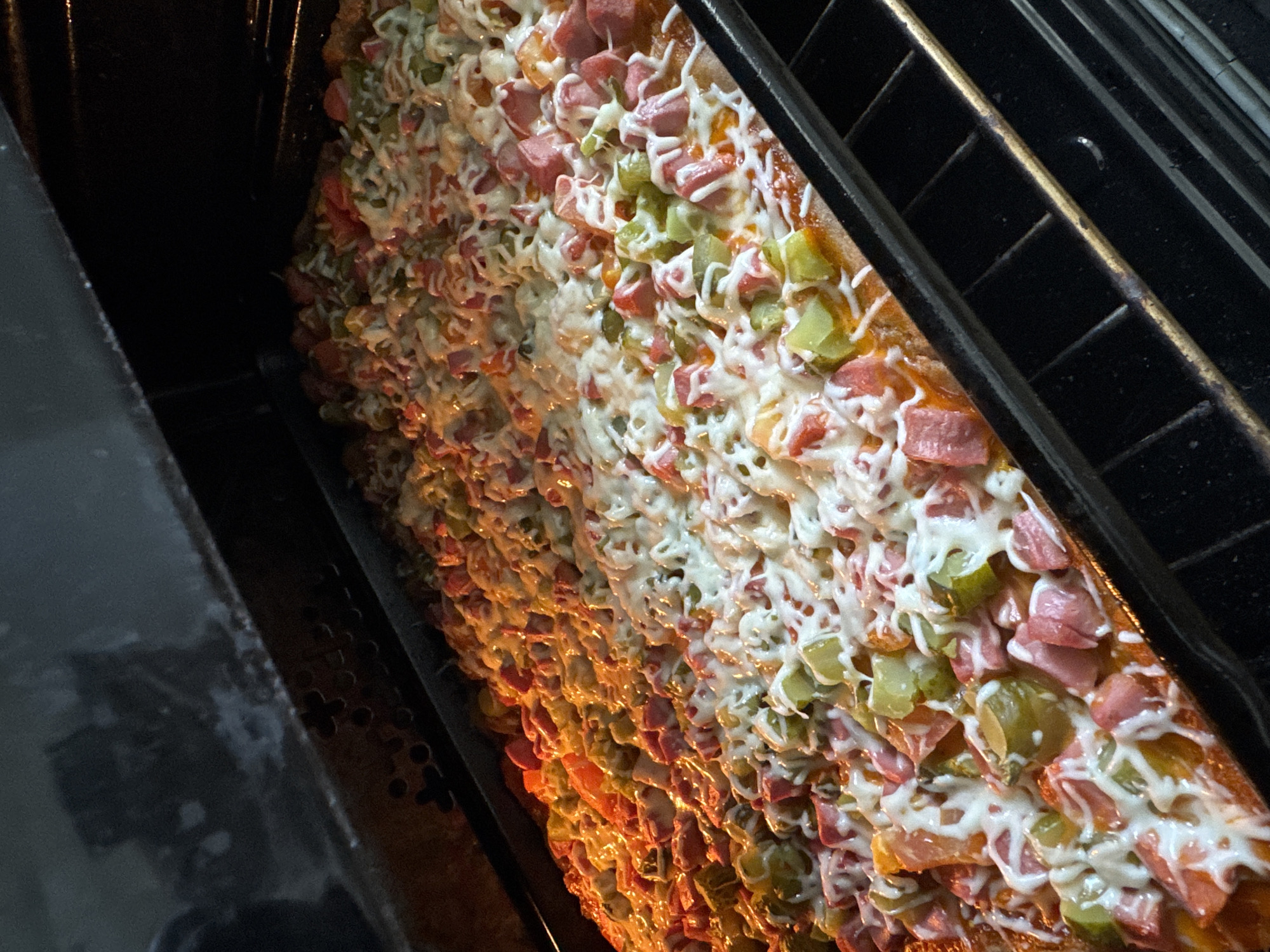 The image shows a rectangular pizza inside an oven, likely in a domestic kitchen setting. The pizza is heavily topped with diced ham, green peppers, and a drizzled white sauce, possibly mayonnaise. The oven appears to be in operation, with visible light and heat affecting the appearance of the pizza. There is no verifiable evidence connecting the image to the city of Chaikovsky, Russia.