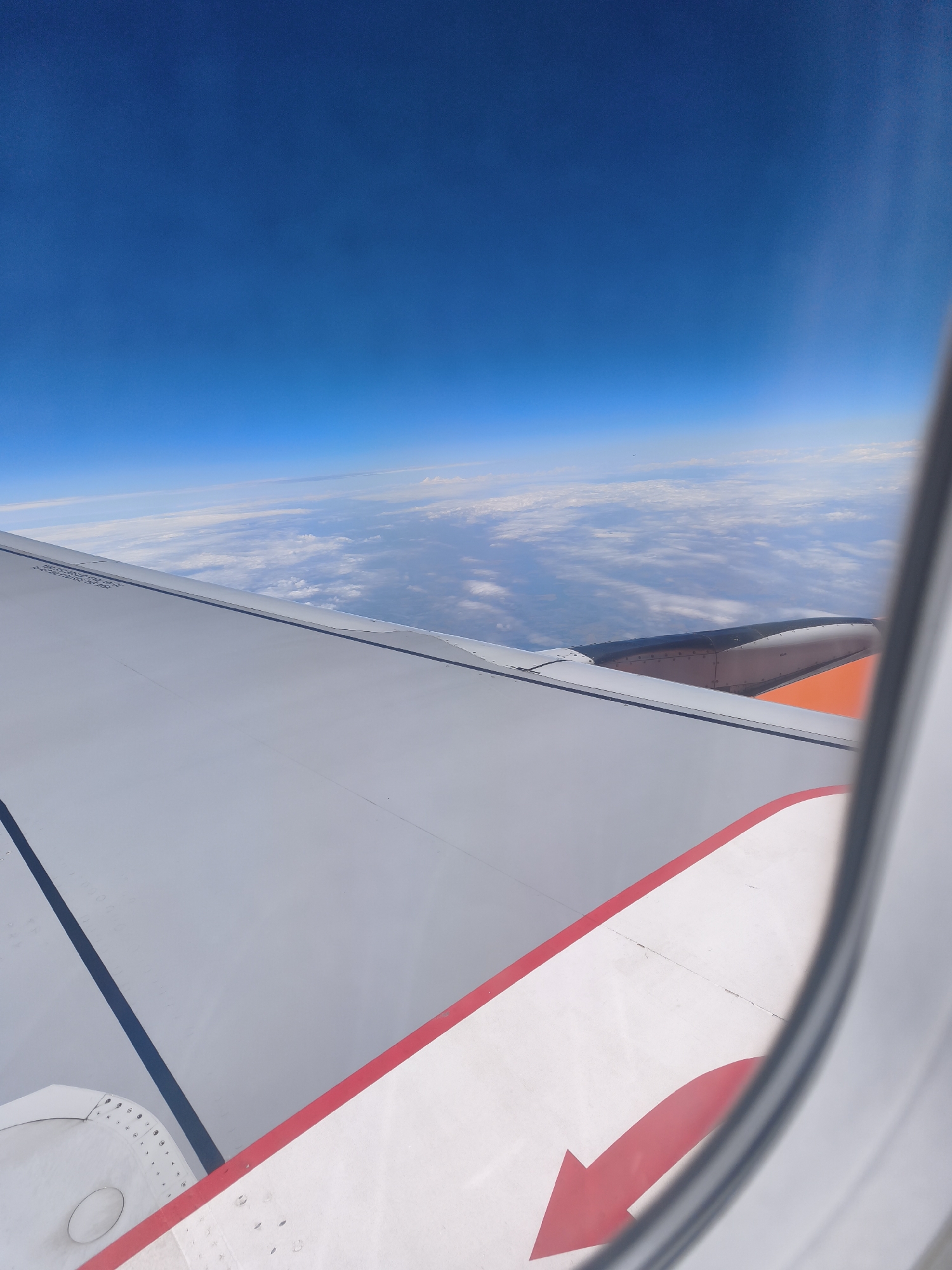 The image shows a view from an airplane window looking out at the sky and clouds. The wing of the airplane is visible in the foreground, with a section painted white with a red arrow pointing to the right. The sky is a clear blue, and there are white clouds below the airplane. The scene is a typical aerial view from a commercial flight. The location in the city of Trebbin, Germany is not determinable from the provided image.