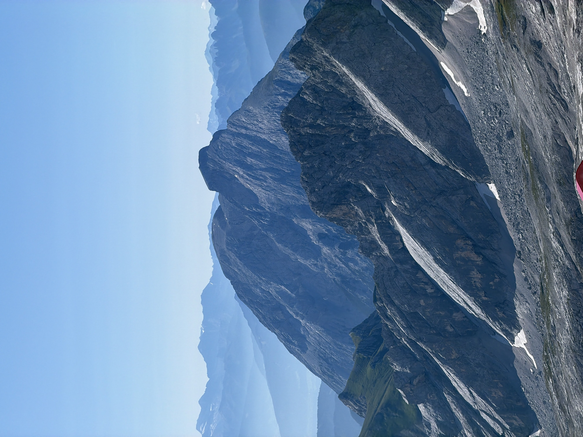 The image shows a vast mountain landscape with multiple layers of peaks fading into the distance under a clear, pale blue sky. The foreground features rugged, steep mountain slopes with rocky scree and patches of snow. The mountains are predominantly grey and dark blue, with some areas of green vegetation on the lower slopes. In the very bottom center of the image, a small, bright red structure, likely a building or shelter, is visible. The scene evokes a sense of height and remoteness, characteristic of alpine environments. The image appears to be a panoramic view taken from a high vantage point.
