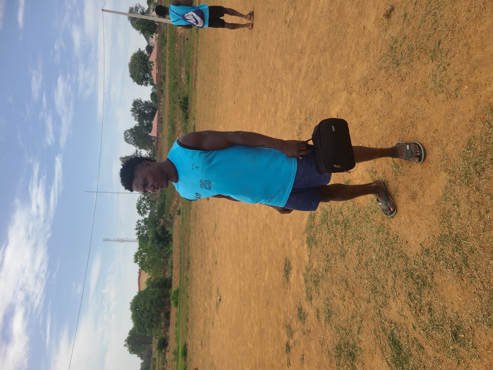 A man wearing a blue athletic tank top and dark shorts is standing on a field with sparse grass, holding a black bag. He is wearing flip-flops. In the background, another person wearing a similar top is walking away. A transmission tower is visible in the background. The scene is in Barikin Jirga Summa, Nigeria. The ground is a dry, brownish color. There are trees lining the perimeter of the field. The sky is blue with scattered clouds.