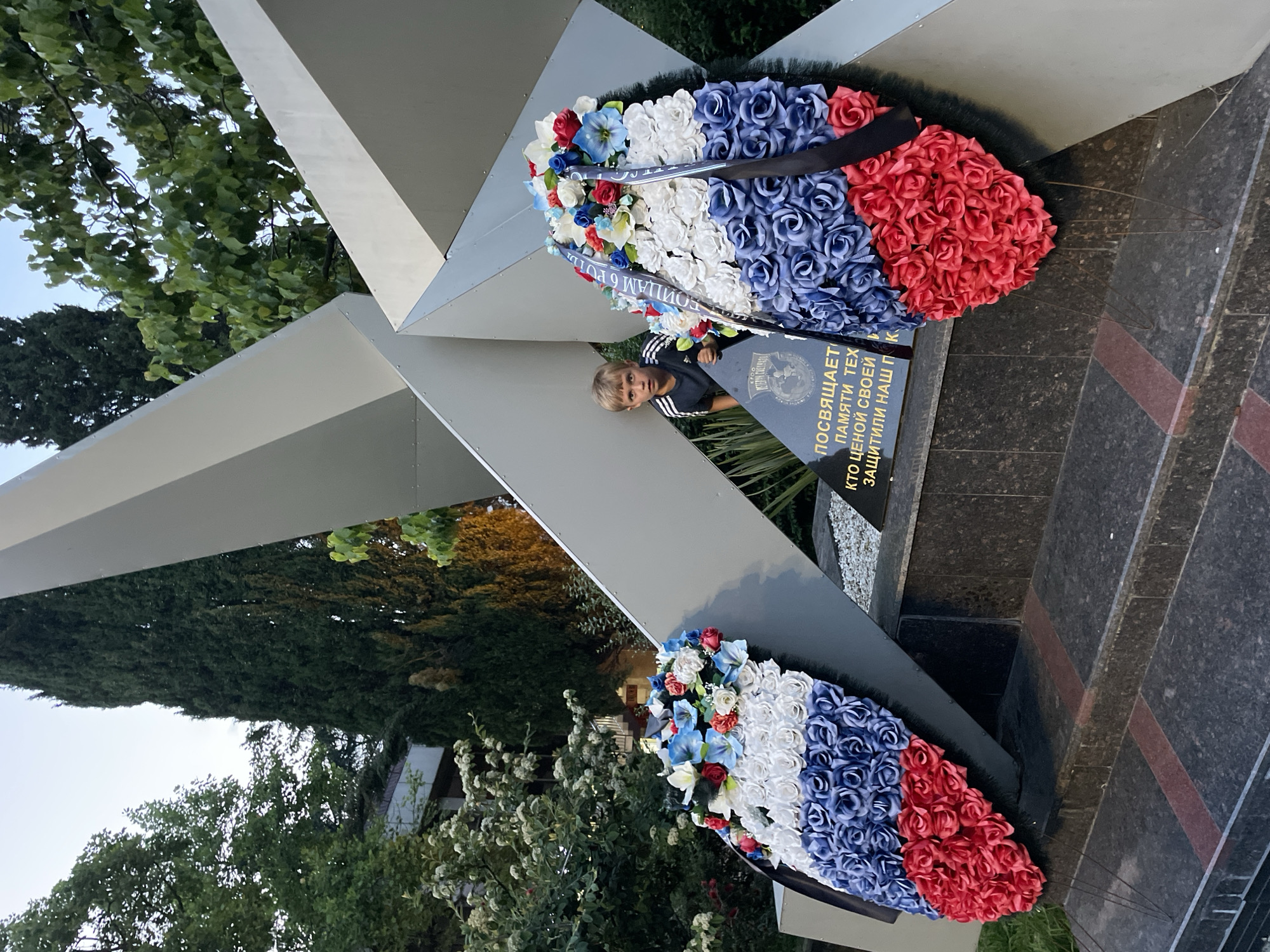 A young boy with blonde hair stands behind a memorial in Sochi, Russia. The memorial features a large, stylized silver star with sharp angles, behind which the boy is partially visible. Two large wreaths, adorned with red, white, and blue artificial roses, are placed at the base of the memorial. These wreaths mimic the colors of the Russian flag. A black plaque with text in Russian is visible at the base of the memorial, between the wreaths. The surrounding environment includes lush green trees and foliage, as well as a building in the background. The sky above is clear and light blue, suggesting it is either early morning or late afternoon. The overall scene appears to be a solemn or commemorative one, given the presence of the memorial and wreaths.
