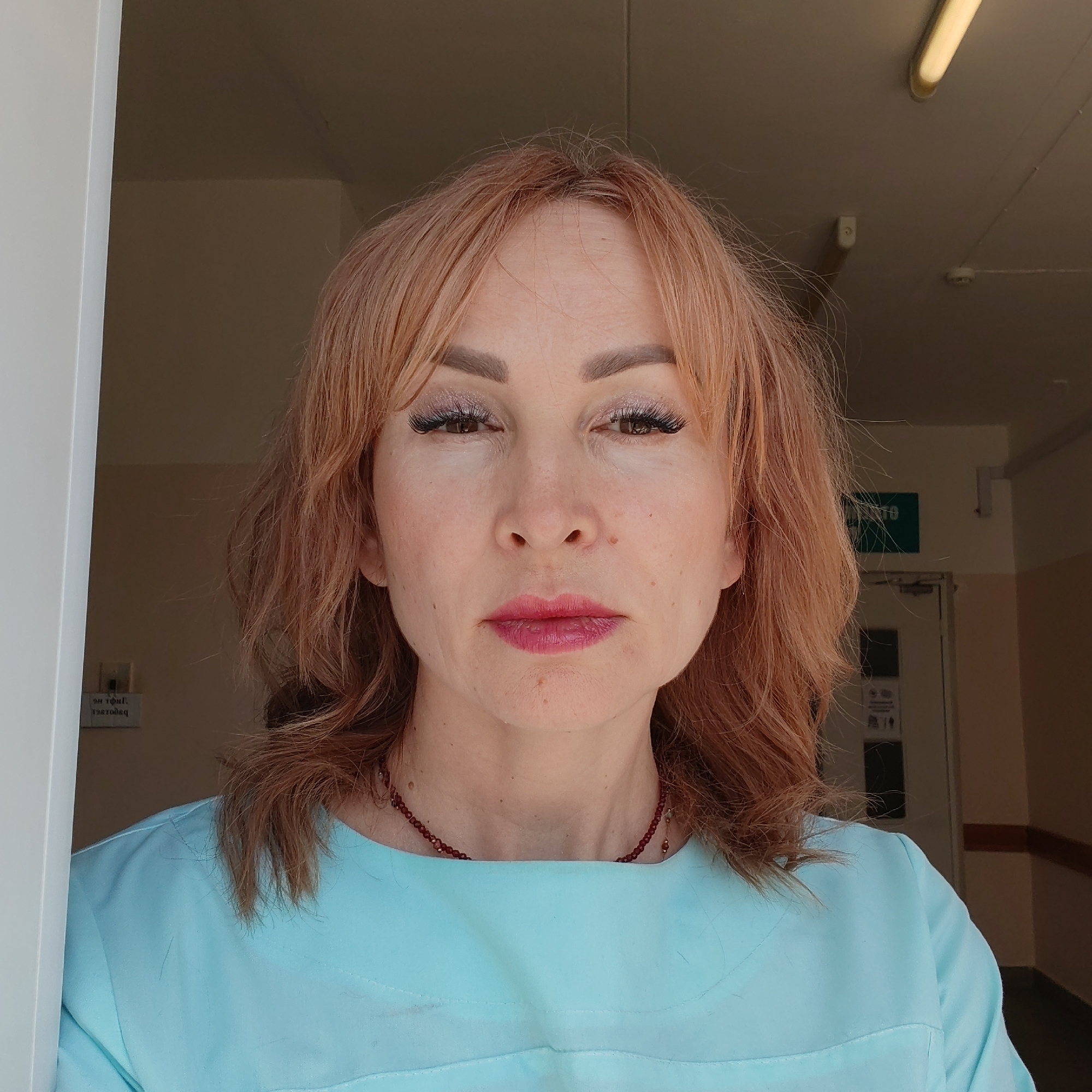 A middle-aged woman with reddish-blonde hair is centered in the frame, looking directly at the viewer. She is wearing a light blue top and a dark red beaded necklace. She has makeup on her eyes and lips. The background shows a doorway or entrance to a building. To the right, a green sign is visible above a door. The location is likely within a building in Chaikovsky, Russia.