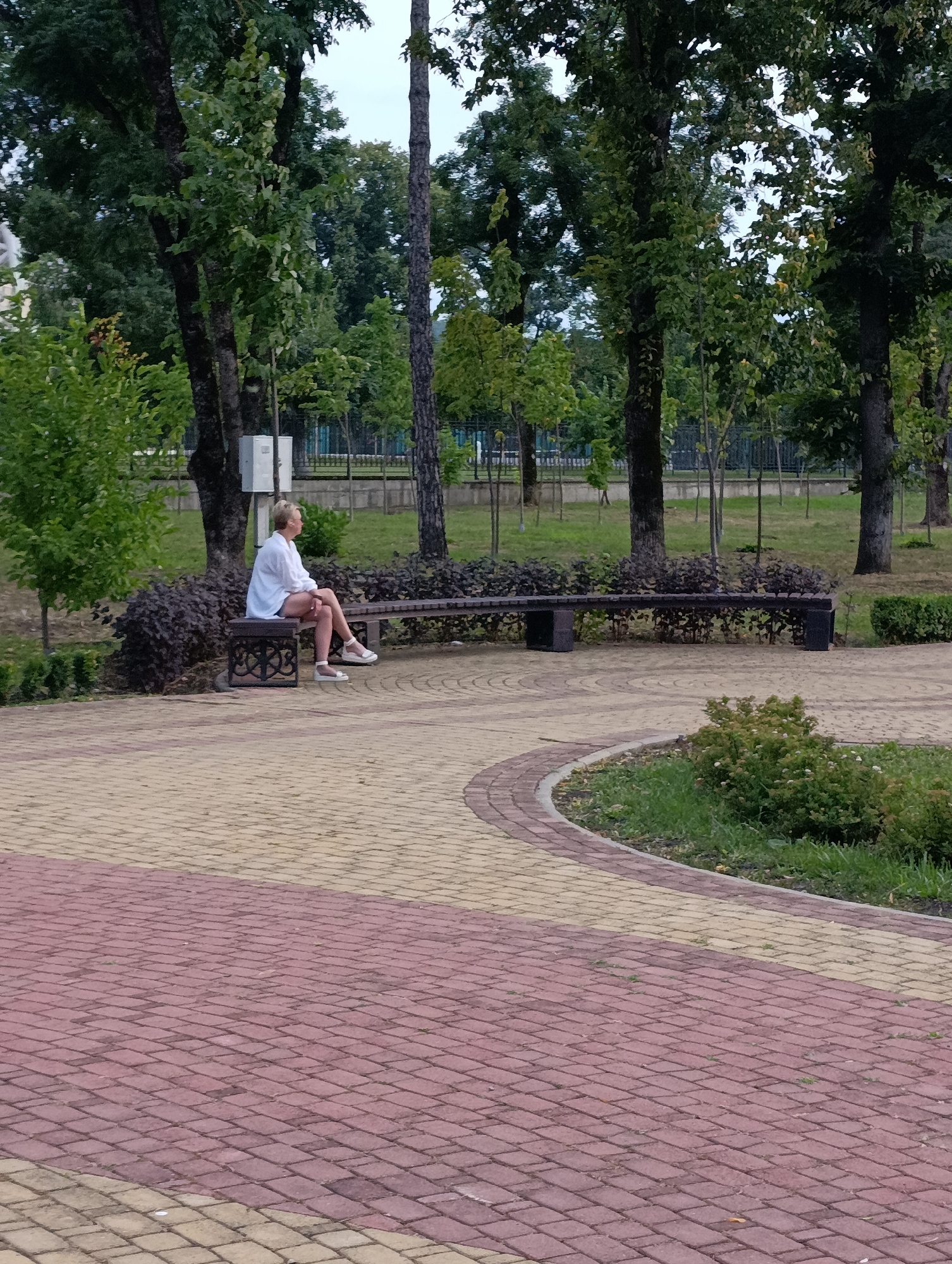 A woman with blonde hair is sitting on a park bench, facing right and looking away from the camera. She is wearing a white shirt and dark shorts. The bench is located in a park with a paved walkway in the foreground. The walkway is made of red and yellow bricks in a pattern. Behind the bench, there are trees and bushes, as well as a fence. The sky is overcast. The image was taken in Maykop, Russia.