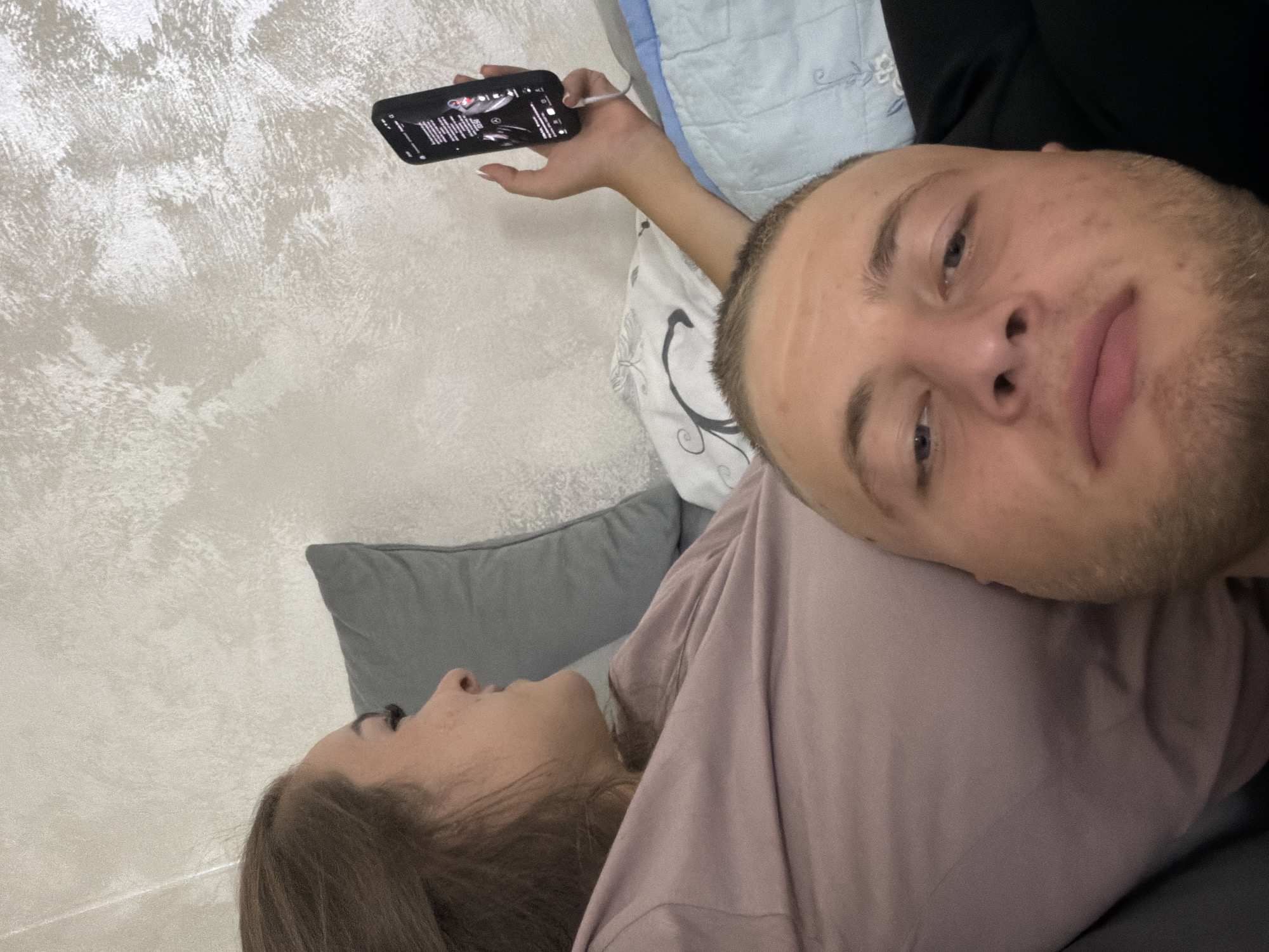 A close-up, first-person perspective shot shows two individuals in what appears to be a bedroom. The camera is positioned low, focusing on the face of a man with light skin, short blonde hair, and blue eyes, who is looking directly at the camera. His mouth is slightly open, and he has stubble on his chin and cheeks. To his left and slightly behind him, a woman with brown hair is visible in profile. She is wearing a mauve-colored top and has her head resting on a grey pillow. Her eyes are directed towards a smartphone she is holding in her right hand, which displays a black screen with white text and what appears to be a song or video playing. A white cable connects the phone to an unseen device. The background consists of a textured, light-colored wall and a blue and white patterned blanket or duvet. The scene suggests a moment of relaxation or shared entertainment. The location is specified as being in the city of Strășeni, Moldova.