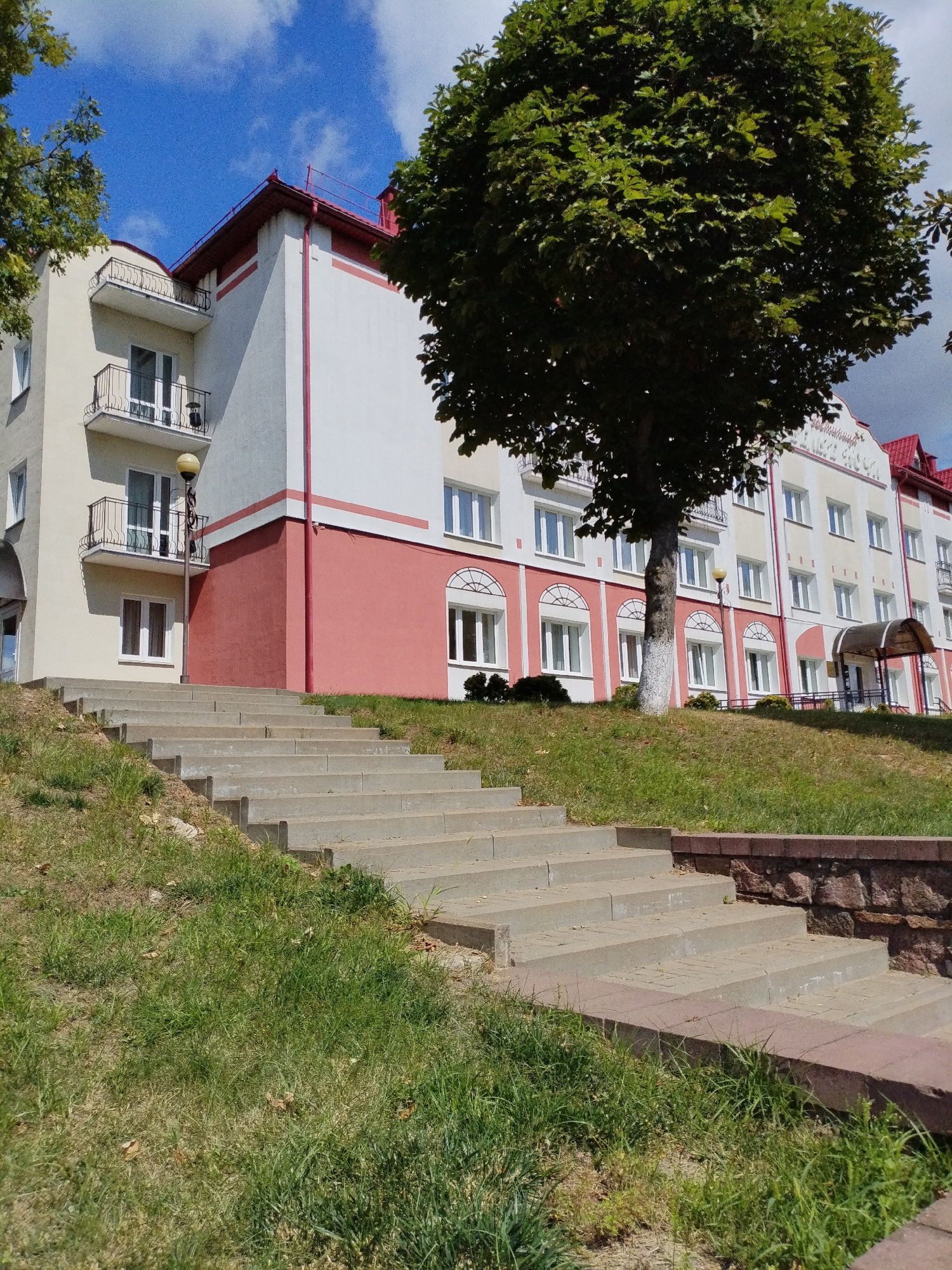 The image captures a view of a multi-story building with a red roof and white and pink facade in Bialynichy, Belarus. A large, leafy green tree stands prominently in front of the building, partially obscuring it. In the foreground, a wide, concrete staircase ascends a grassy incline. The sky is blue with scattered white clouds, suggesting it is daytime. The building appears to be an apartment complex or a public institution, characterized by numerous windows and balconies. A small covered entrance with a sign is visible towards the right side of the building. The overall scene depicts an outdoor urban setting with a blend of architectural and natural elements.