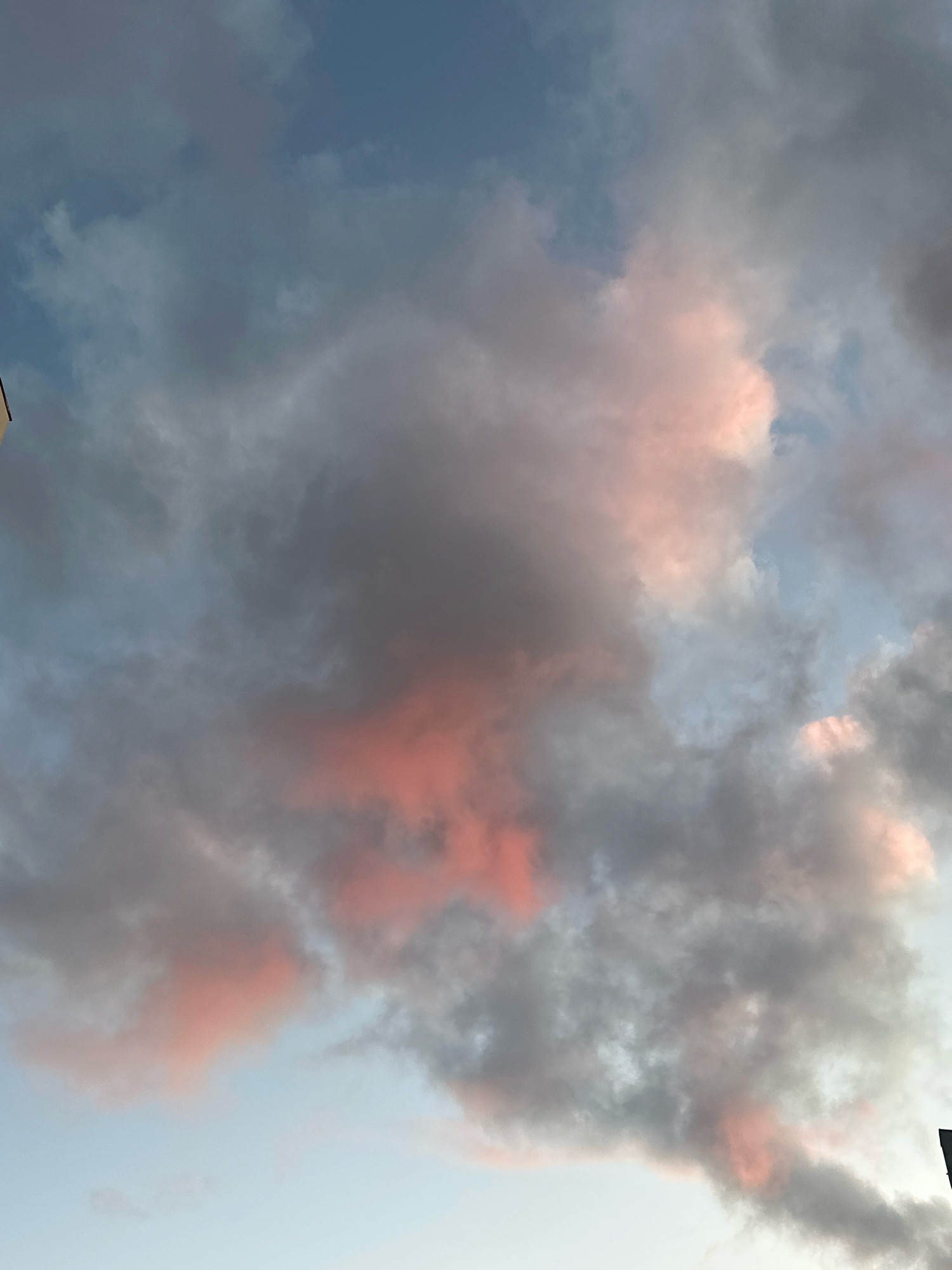 The image displays a sky filled with a mix of gray and pinkish clouds. The lighting suggests it is either sunrise or sunset. The bottom right corner of the image shows a sliver of a dark, vertical object, possibly a lamppost or part of a building. There is no indication of the city of Minsk, Belarus, or any specific geographical features. The scene is entirely focused on the sky and clouds.