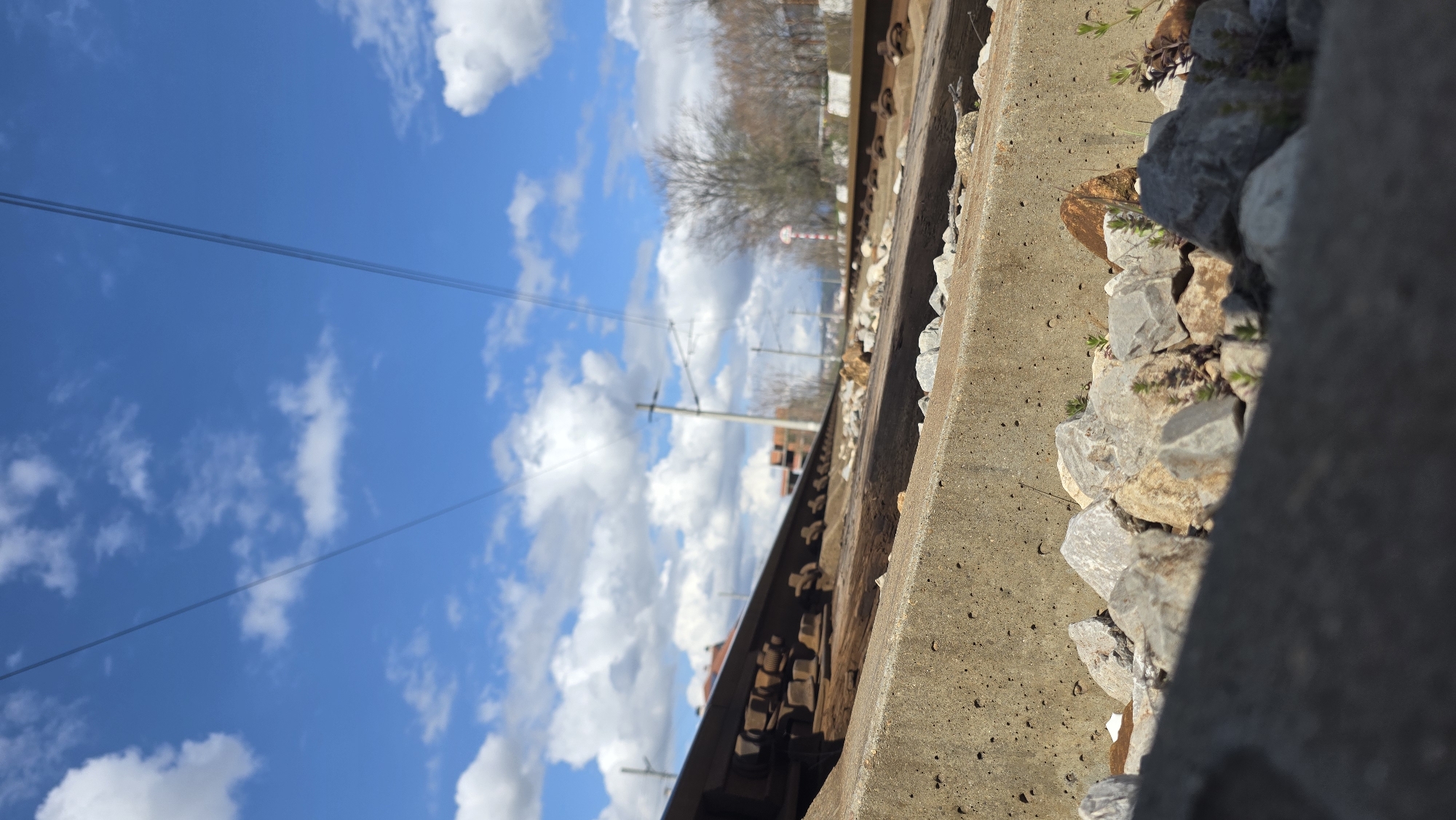 The image shows a close-up, low-angle view of a railway track and the surrounding environment, likely taken near Blagoevgrad, Bulgaria. The sky is visible, filled with white clouds and a clear blue background. Electrical wires run across the sky. The railway tracks are rusted and heavily textured. The concrete base of the track is edged with gravel and small rocks. In the background are trees, and what appears to be a red and white striped railway sign.