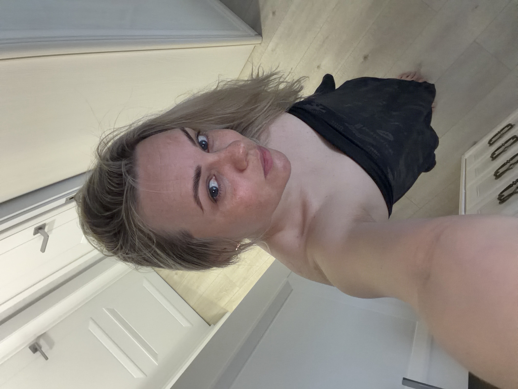 A woman with blonde hair and blue eyes is taking a selfie in a brightly lit room. She is wearing a dark garment over her shoulder and chest. The room appears to be a bathroom with white cabinets and drawers visible in the background. The floor is light-colored wood. There is no indication of the location being Votkinsk, Russia.