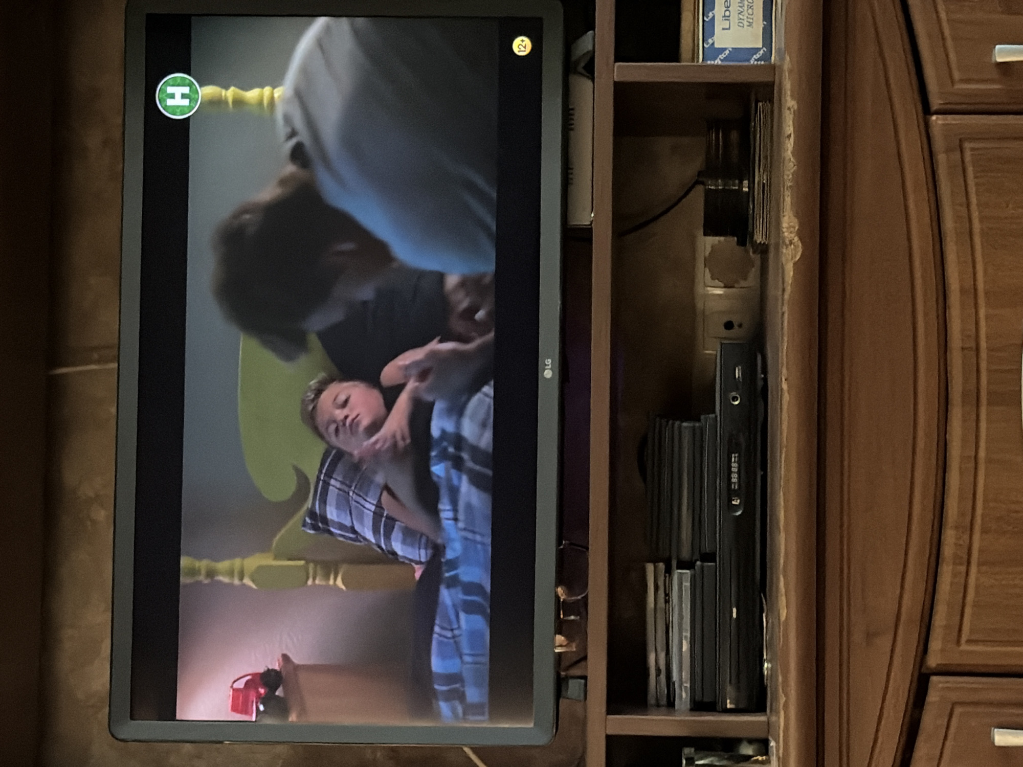 The image shows a television screen displaying a scene of two people, one adult and one child, in a bedroom. The child is lying on a bed with blue and white plaid bedding, and an adult is leaning over them. A red toy car is on a bedside table. A green circular logo with an "H" in white is visible in the top right corner of the screen. A "12+" rating symbol is in the bottom right corner. The television is positioned on a wooden shelf unit, which also holds stacks of DVDs or CDs, a DVD player, and other electronic devices. A blue and white box with text is on the right side of the shelf. The room appears to be a domestic setting.