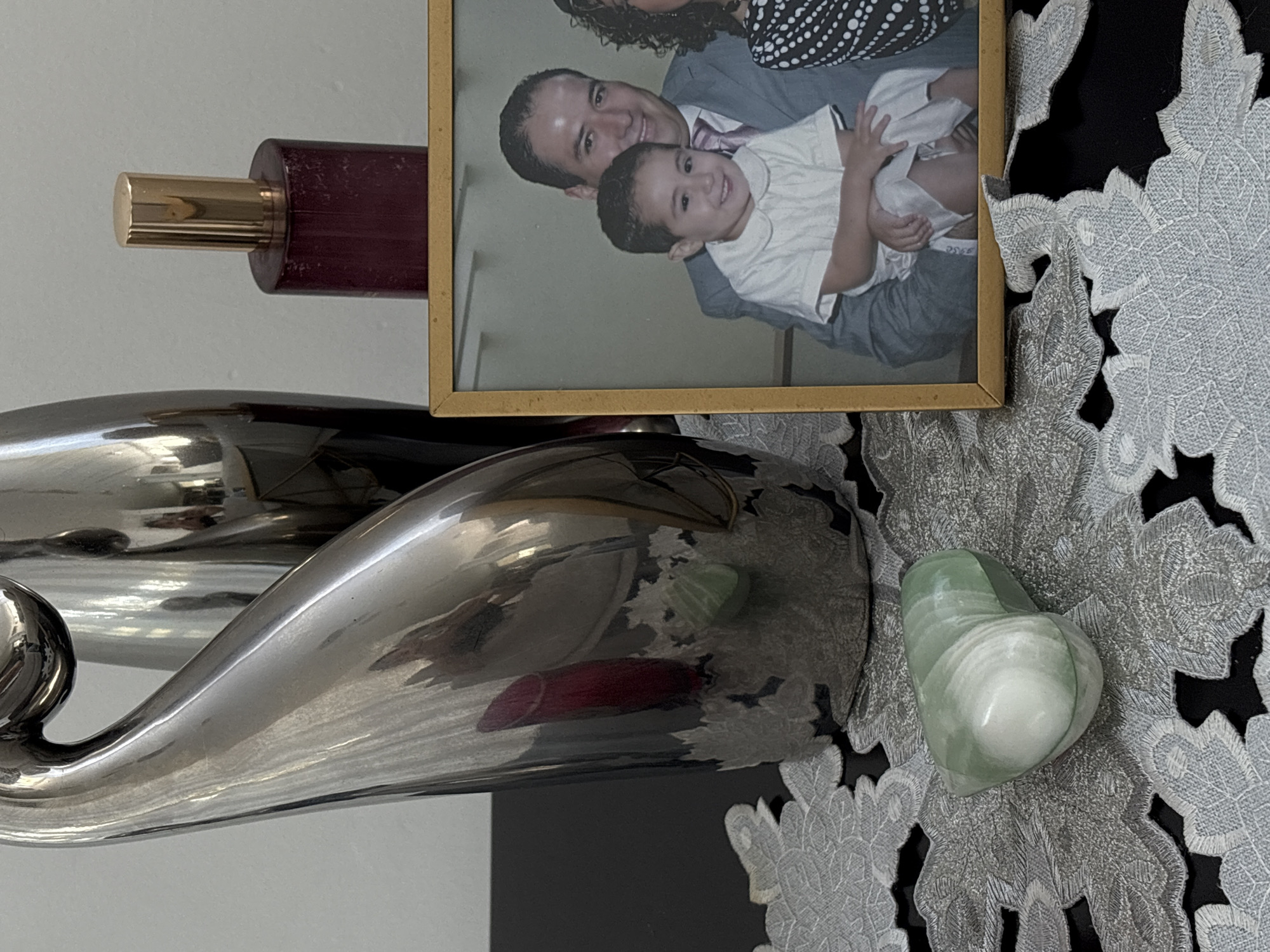 This image, set in San Bernardino Tlaxcalancingo, Mexico, captures a close-up of a decorative arrangement on a surface.
The primary subject is a gold-framed photograph, prominently displayed in the upper right quadrant of the image. The photo depicts a family portrait, with a man smiling broadly while holding a young child, who is also smiling. The man wears a light-colored suit and a tie, while the child is dressed in a white collared shirt. Part of another adult, with curly dark hair, is visible behind the man's head. All individuals in the photograph appear joyful.
To the left of the framed photo, a dark red, cylindrical bottle with a metallic gold cap stands upright, possibly a perfume or a decorative container. Below these items, a large, highly reflective, silver-colored decorative object with a sleek, organic, and wavy design dominates the lower and central left portions of the image. Its polished surface shows distorted reflections of the surrounding objects, including a reddish hue from the bottle and a green tint, possibly from the heart-shaped stone.
On the right side of the image, resting on a decorative textile, is a light green and white heart-shaped stone, exhibiting natural striations in its material. The entire display rests on a grey or silver-colored lace placemat or tablecloth, which features intricate floral and leaf patterns against a darker background.
No specific activity is taking place in the immediate scene, which is a still life arrangement. The photograph captures a happy family moment. There is no visible text in the image.
