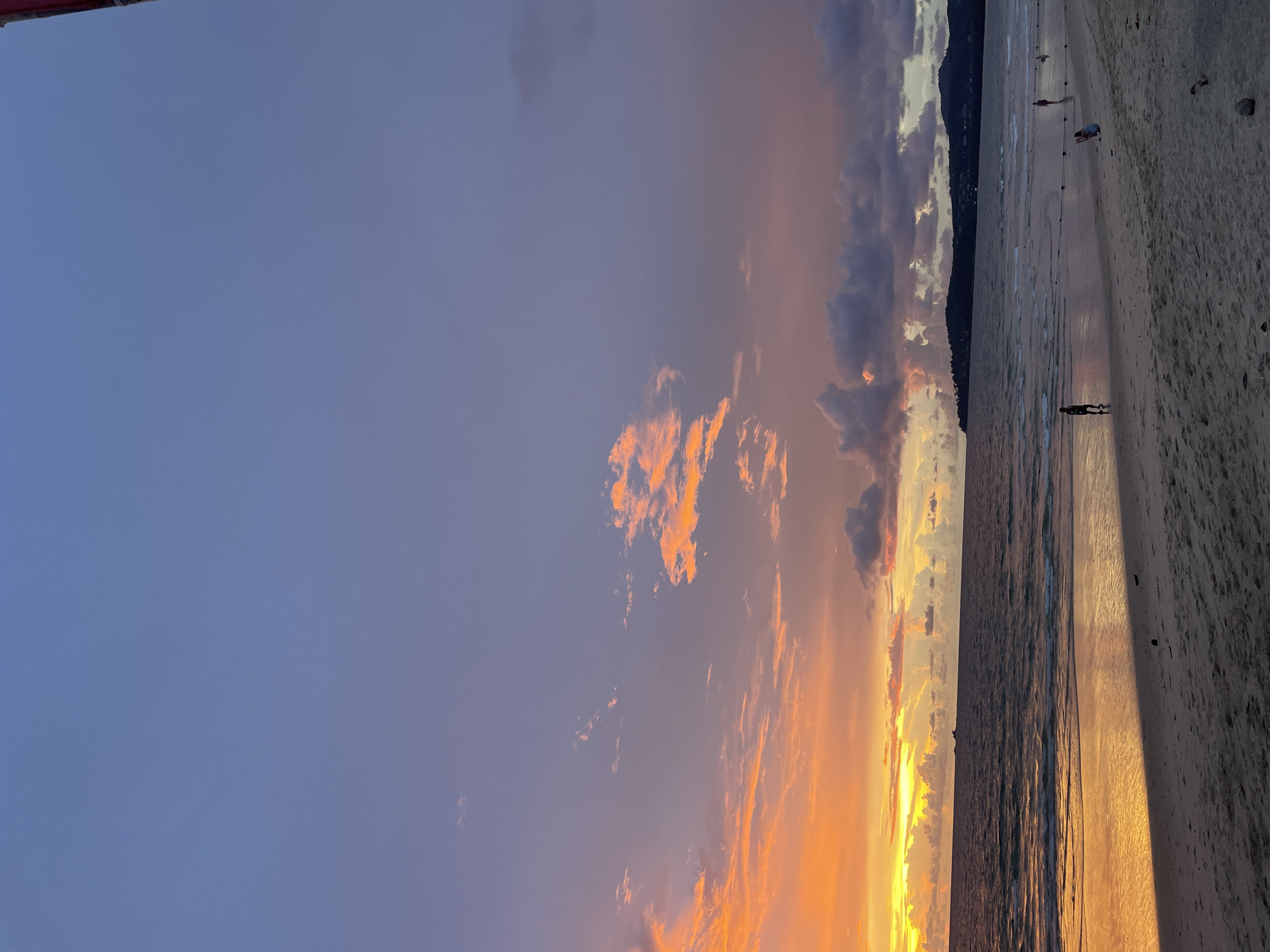 This is a photograph of a sunset at a beach in Choeng Thale, Thailand. The sky is a gradient of purple, pink, and orange, with scattered clouds. The ocean is dark and reflects the colors of the sky. The beach is sandy, and there are a few people walking and standing on it. The overall impression is one of tranquility and beauty.