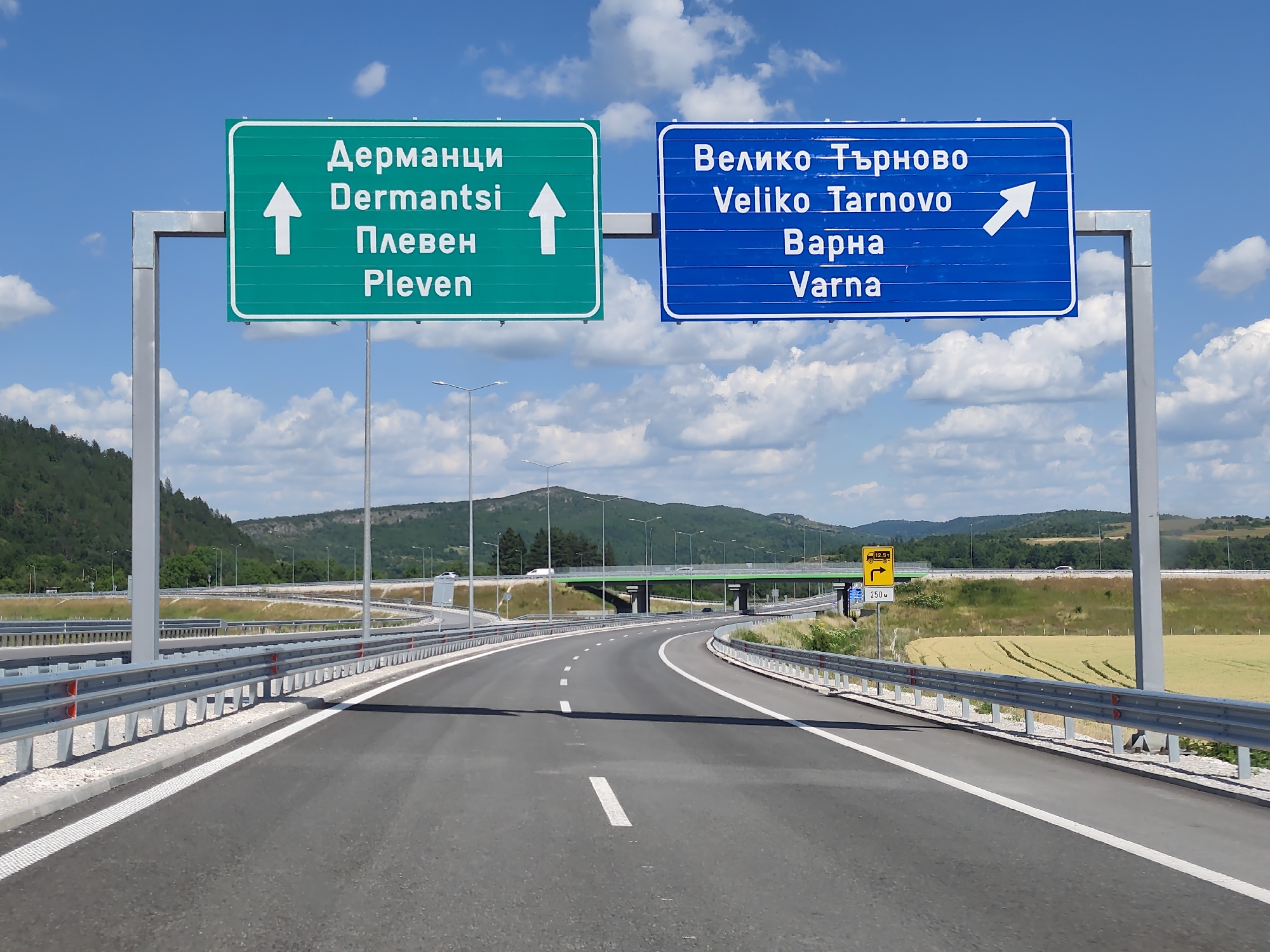 A bright, sunny day in Brestnitsa, Bulgaria, as seen from a highway. The sky is a clear blue with scattered fluffy white clouds. The scene features a modern highway with two lanes in each direction, separated by a concrete barrier and guardrails. On the left, a green directional sign indicates "Дерманци Dermantsi" and "Плевен Pleven" with an upward arrow, meaning these destinations are straight ahead. On the right, a blue directional sign displays "Велико Търново Veliko Tarnovo" and "Варна Varna" with an arrow pointing to the right, indicating these cities are that way.
In the background, rolling green hills covered with trees are visible. Beyond the highway on the right, there's a field of golden wheat, with tire tracks suggesting recent activity. An overpass, painted green, crosses the highway in the distance. Several streetlights line the highway, and other vehicles can be seen on the road and overpass. A smaller yellow sign is visible on the right, indicating a weight limit of 12.5 tons for trucks and a distance of 250 meters to a turn. The overall impression is one of a well-maintained, modern road infrastructure in a scenic, rural Bulgarian setting.