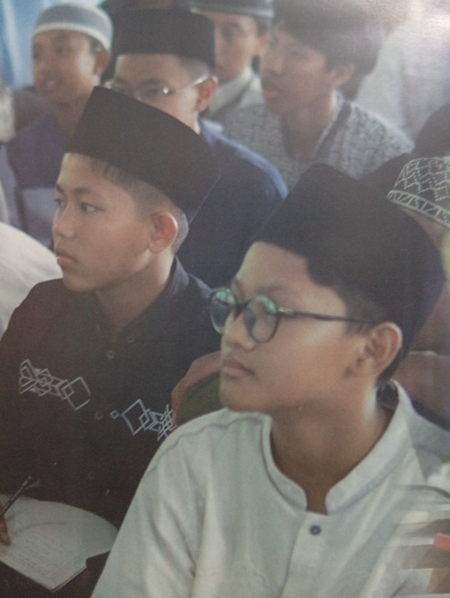This image depicts a group of young males, likely students, wearing traditional Islamic attire. Two individuals are prominently featured in the foreground, both wearing black peci hats. The individual on the left is looking to the left of the frame, with a serious expression. They are wearing a dark collared shirt with a white geometric pattern on the chest. To their right, another young male is wearing glasses and looking forward. They are dressed in a light-colored collared shirt. In the background, other individuals are partially visible, also wearing peci hats and traditional clothing. The setting appears to be an indoor gathering or event. Without further information or visual cues within the image itself, it is not possible to definitively confirm the location as Tlogomulyo, Indonesia.