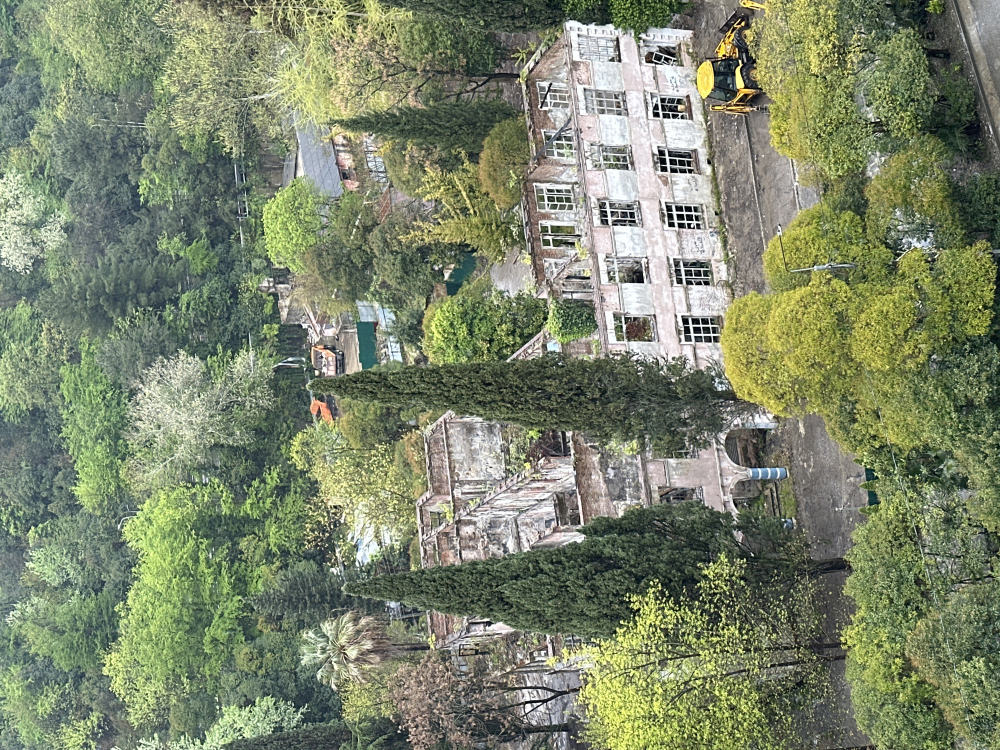 The image shows a partially demolished building in Sochi, Russia, surrounded by lush greenery. The building has multiple floors with missing windows and sections of walls, indicating significant damage. A yellow excavator is parked near the top of the building, suggesting ongoing demolition work. The surrounding environment consists of dense trees and vegetation, indicative of a forested or park-like setting. There are other structures visible in the background, partially obscured by trees. A few blue pipes can be found near the building.