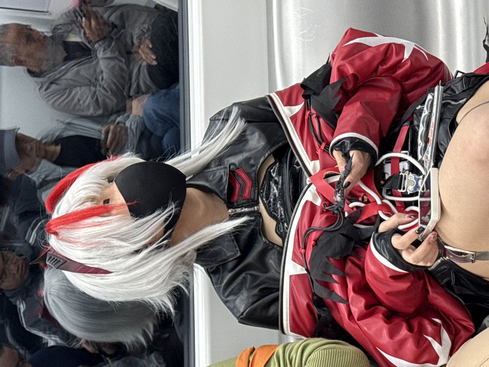 The image shows a person in a black and red costume, possibly a cosplay outfit, with white hair streaked with red. They are wearing a black bra-like top and a black leather jacket with red and white accents. The jacket is overlaid with a red garment with white star-like patterns and black decorative elements. The person is also wearing black gloves with long, pointed fingernails. A silver belt-like accessory is visible around their waist.
The person is reflected in a mirror or glass surface, revealing a view of a room. In the reflection, an older man wearing a grey jacket and blue jeans is visible, along with other indistinct figures. The man appears to be observing or possibly taking a photo of the person in the costume.
The environment suggests an interior space with neutral-colored walls. There is no information to pinpoint this image to Liangjiang, China.