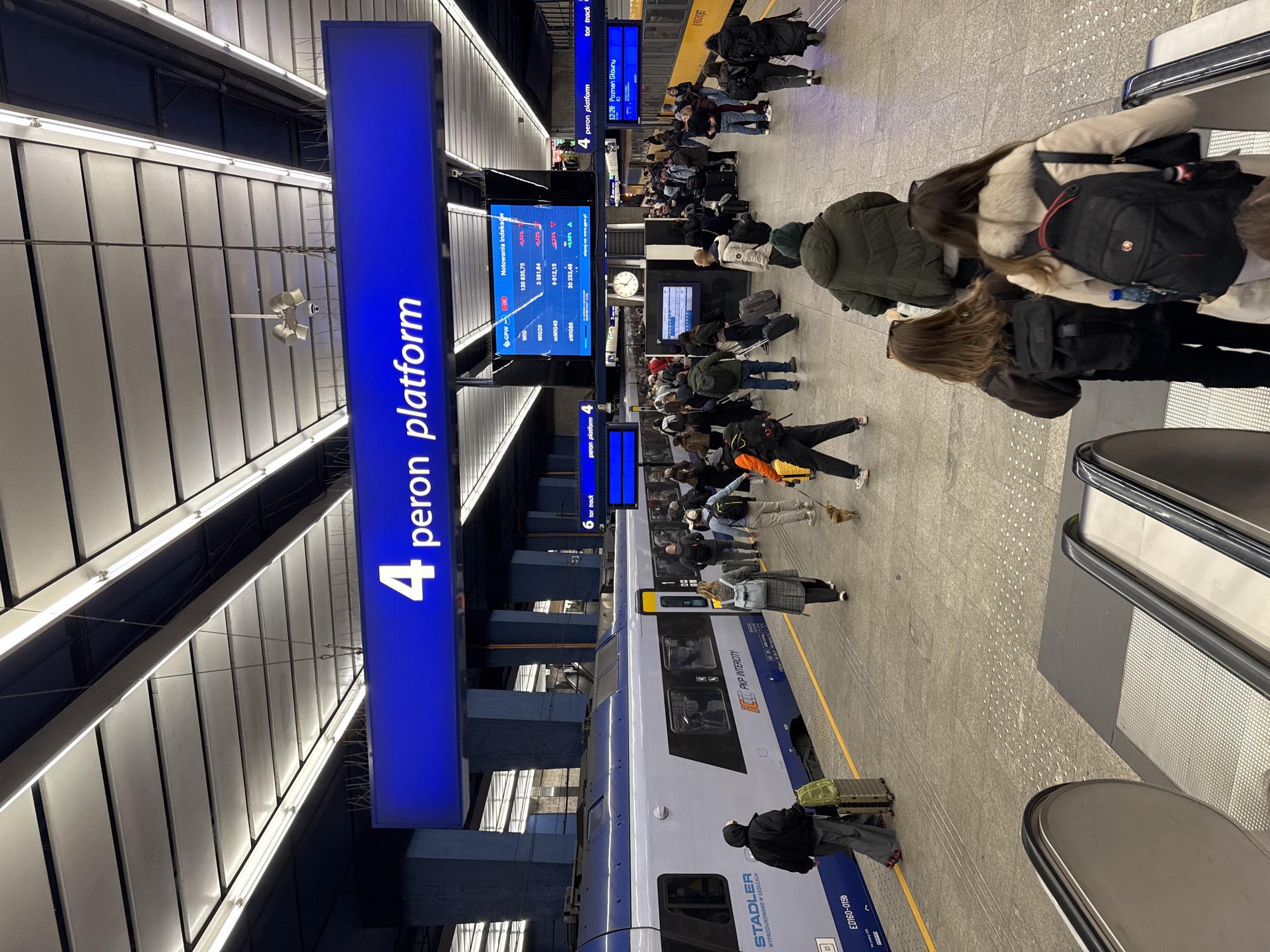 The image depicts a bustling scene at a train station platform, likely in Warsaw, Poland, based on the visible signage and train design. The viewpoint is from a high-angle perspective at platform 4. A blue sign indicates "4 peron platform." Passengers with luggage wait near a modern white and blue PKP Intercity train manufactured by Stadler. Information screens display train schedules and financial data. A yellow line runs along the platform edge. People are near an escalator.