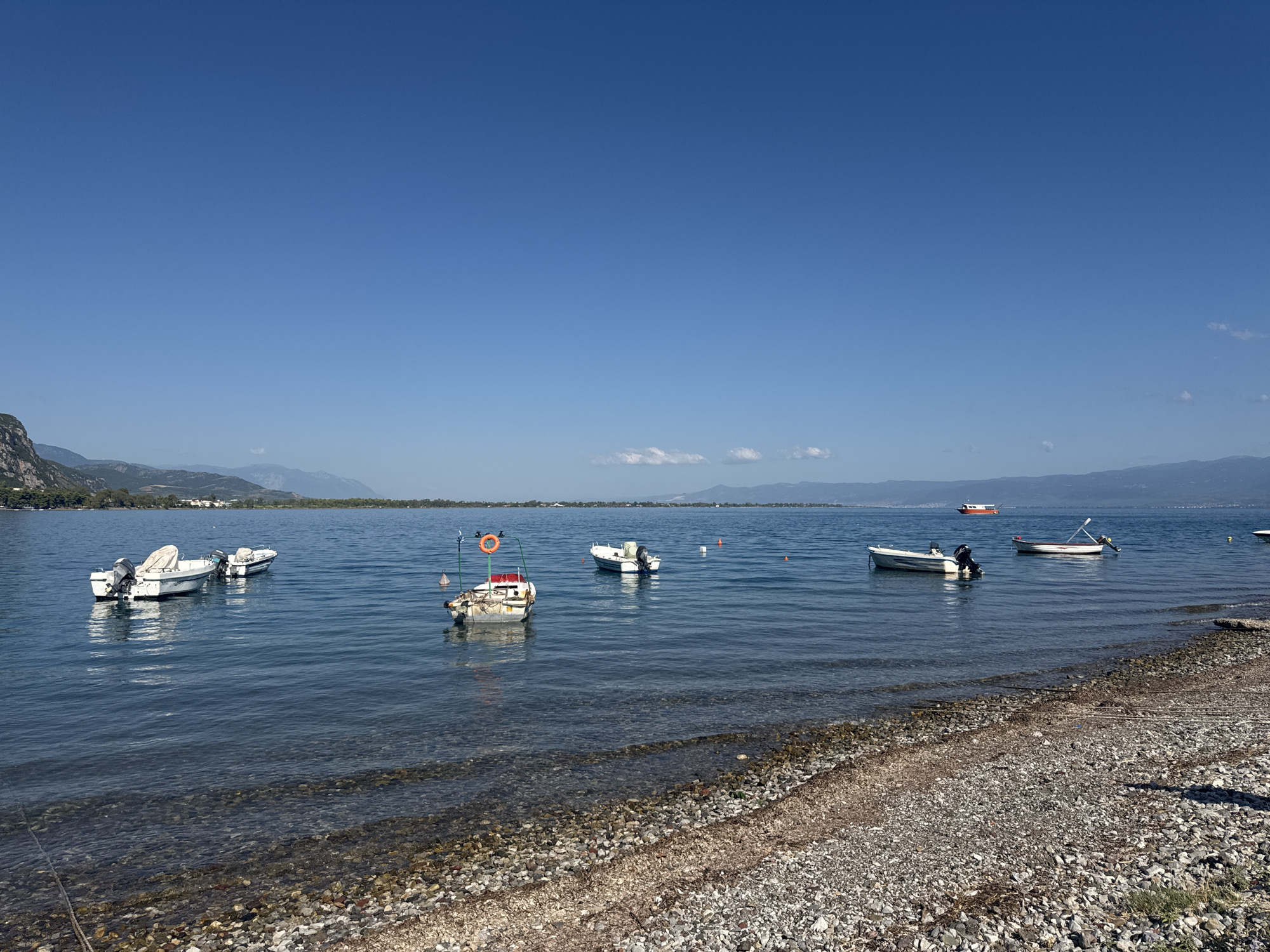 The image shows a body of water, likely a bay or lake, with several small boats anchored or moored near the shore. The water is calm and reflects the clear blue sky. The shoreline is composed of pebbles and a small amount of vegetation. In the background, there are rolling hills and mountains under a cloudless sky. The scene appears to be a tranquil coastal or lakeside setting. Based on the scenery, the location is likely the Municipal Unit of Kamena Vourla, Greece, known for its coastal geography and mountainous backdrop.