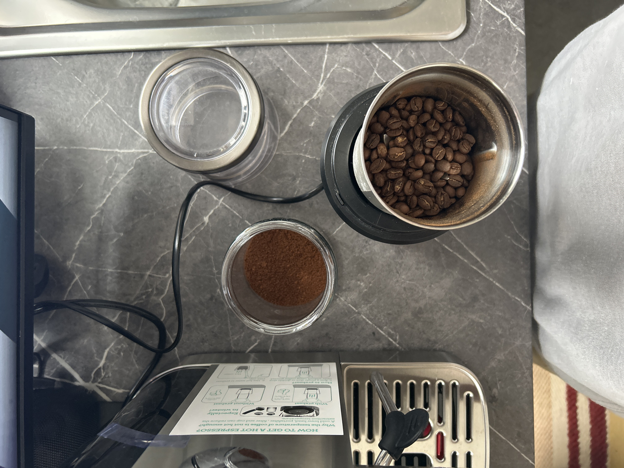 Here is a detailed description of the image:
**Scene:** A high-angle shot showing a countertop scene with coffee-related items.
**Objects:**
* **Coffee Beans:** A metal container is filled with roasted coffee beans.
* **Ground Coffee:** A clear glass container with a silver lid holds ground coffee.
* **Empty Container:** Another clear glass container with a silver lid appears to be empty.
* **Coffee Grinder:** A coffee grinder is partially visible with coffee beans in it.
* **Espresso Machine:** Part of a silver espresso machine is visible at the bottom of the frame, along with an attached instruction manual.
* **Countertop:** The objects rest on a gray countertop with a marble-like pattern.
**People:**
* A portion of a person is visible on the upper right with a part of the torso wearing a white garment.
**Environment:**
*The counter seems to be set up for making espresso.
**Location:**
The location is not specified in the picture, so there is no way to know whether this picture was taken in London or elsewhere.