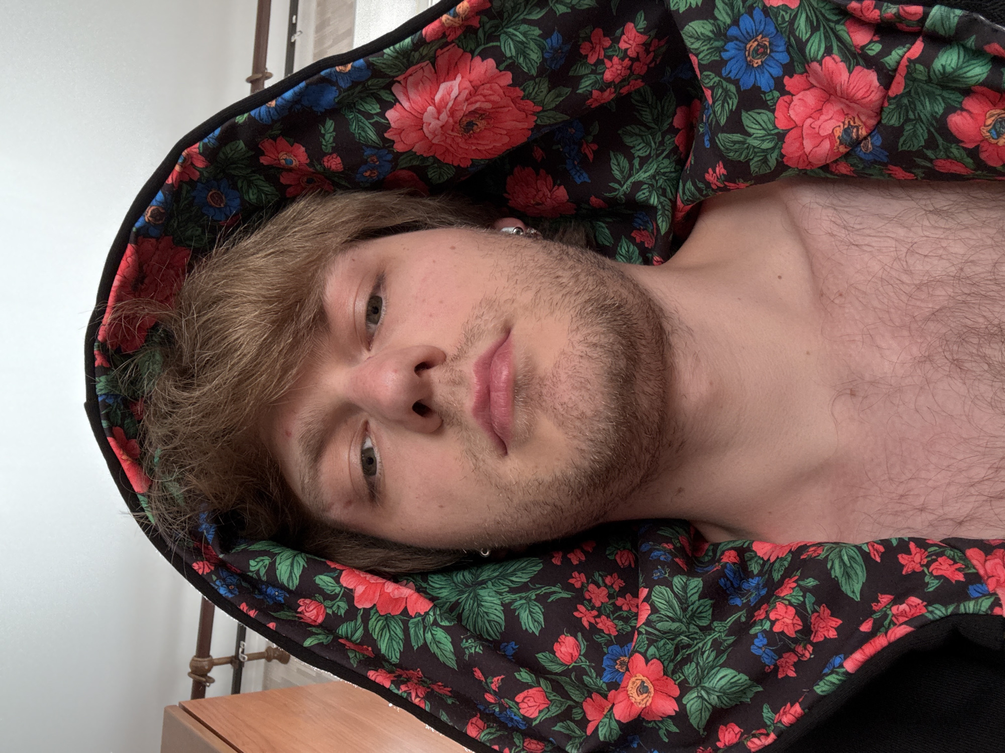 A close-up, angled selfie shows a person lying down with their head resting on a pillow adorned with a vibrant floral pattern. The pattern features large red and pink flowers with green leaves, interspersed with smaller blue flowers. The person has light brown hair, which is slightly messy, and a light beard. They are wearing a black hoodie with the floral pattern lining the hood, framing their face. The person's left ear is visible, and they are wearing a small silver hoop earring. Their skin is fair, and they have a light dusting of chest hair visible. The lighting suggests it might be daytime indoors, as the light source is not directly visible, but there are hints of natural light from a window. The background includes a plain white wall and a wooden dresser, with a metal fixture that appears to be part of a radiator or shelving unit. The person's expression is neutral, with their eyes open, looking towards the camera. The focus is on the person's face and the striking floral pattern of the hoodie lining. The scene appears to be a casual, indoor setting.