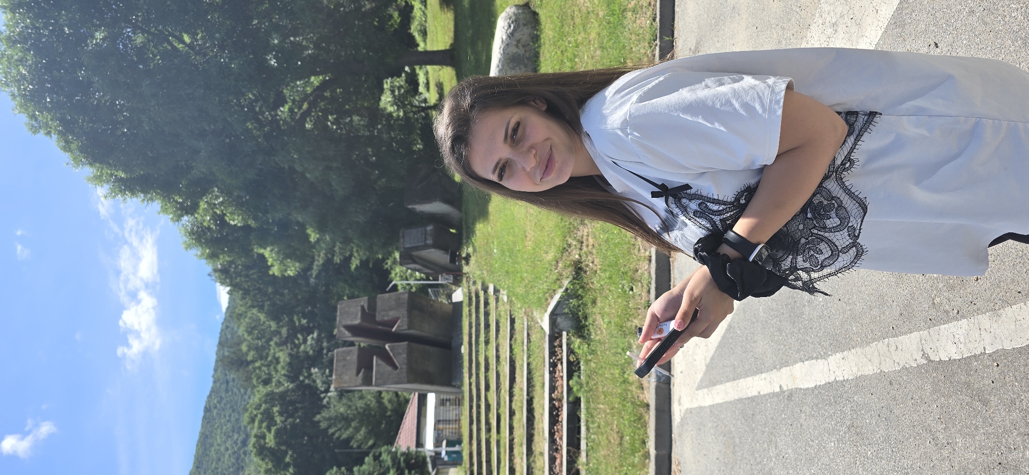 The image features a young woman outdoors under bright daylight, smiling gently at the camera. She has long, dark brown hair and is wearing a light-colored, short-sleeved top with black lace detailing and a small black bow near the neckline. On her left wrist, she wears a black wristband, possibly a smartwatch, and a black scrunchie. Her fingernails are painted light pink.
In her right hand, she holds two small cylindrical objects: a black one, likely an electronic vape pen, with the faint text "UPPO" visible on its side, and a lighter-colored one, possibly a disposable lighter or another vape device, with partial text resembling "fuzztik." A faint wisp of smoke or vapor emanates from the black object, suggesting she is vaping or has just done so.
The background is a mix of natural and man-made elements. To the left, a dense canopy of green trees fills the upper portion of the frame, beneath a bright blue sky with scattered white clouds. In the mid-ground, a sloping grassy area leads to a series of grey, multi-tiered concrete structures. Some of these structures feature abstract, star-like shapes. A building with a reddish-brown roof is partially visible further back, and distant, tree-covered hills are suggested on the horizon. The foreground shows a grey paved surface with a distinct white line, separated from the grass by a concrete curb.
The bright sunlight indicates it is daytime, likely a sunny late morning or afternoon. The setting appears to be a public outdoor space, possibly a memorial park or a similar site given the monumental concrete structures and what might be grave markers.