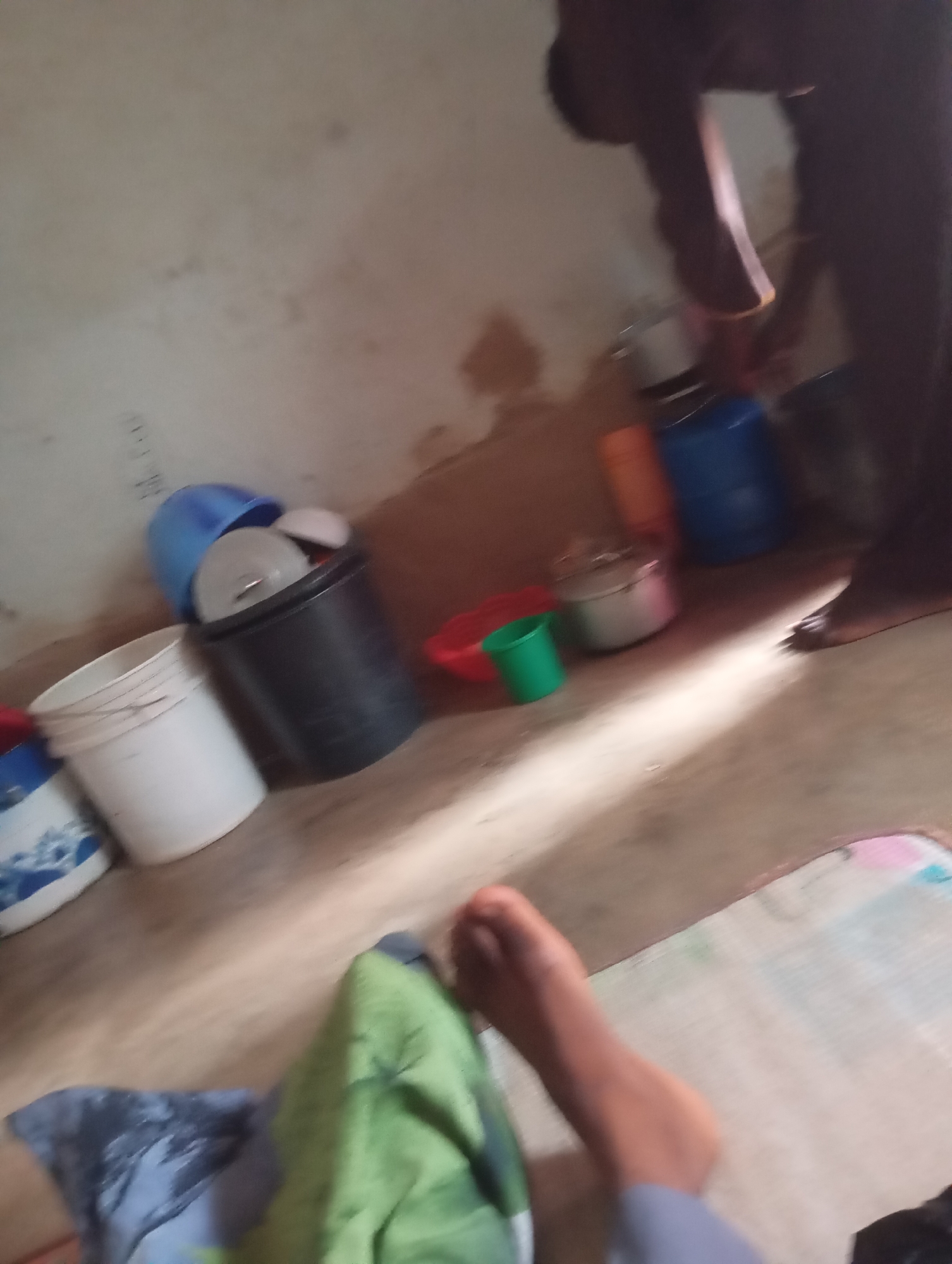 This is a dimly lit indoor scene, likely a room, in Jalingo, Nigeria. The floor appears to be concrete and is sparsely furnished. In the foreground, a person's legs are visible, one resting on a mat that has some faint colorful markings. Beside the legs, a green cloth and a dark blue cloth are laid out.
In the background, there are several objects. A black bucket contains a white lid and a blue bucket is partially nested inside it. To the left, a white bucket is present, and further left, a blue bucket is visible. Near the center, a pot with a lid sits on the floor next to a small green cup and a red bowl. A person, partially obscured by shadow, is standing to the right, appearing to interact with objects on the floor. The wall in the background is rough and discolored. The overall impression is of a utilitarian space with domestic items.