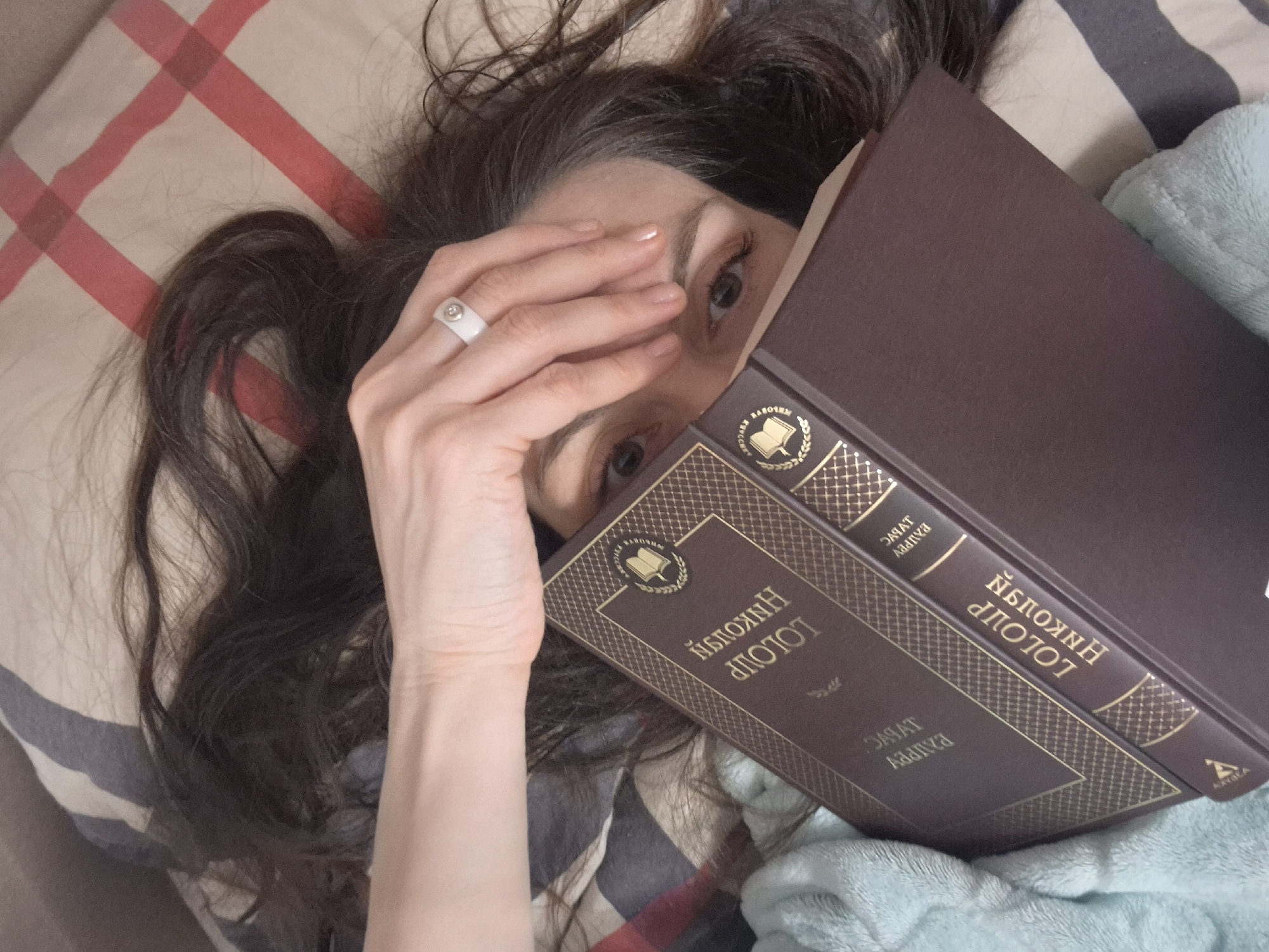 A woman with long brown hair is lying down on a pillow with a plaid pattern, partly covered by a striped blanket, and holding a brown book in front of her face. Her left hand is raised, partially covering her face. The book appears to be titled "Nikolai Gogol." A light blue blanket or soft cloth is partially visible next to the book. The location is Unknown, Unknown.
