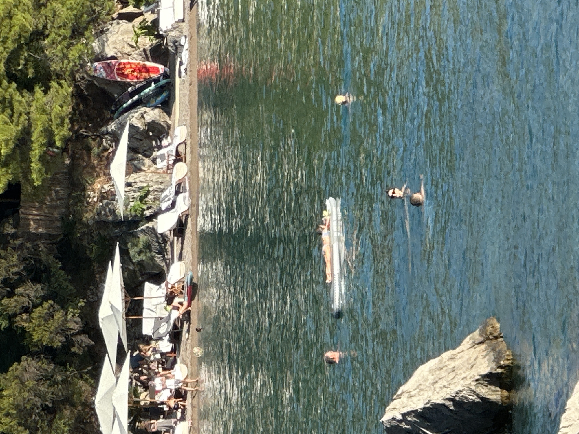 The image depicts a serene scene at a beachside location in Budva, Montenegro. In the foreground, the clear blue waters of the Adriatic Sea gently lap against the shore, with a few rocks emerging from the water on the left side. Several people are enjoying the water. One person is relaxing on a floating inflatable mattress, basking in the sun. Two other individuals are swimming nearby, their heads visible above the water.
The middle ground features a sandy beach lined with lounge chairs and large white umbrellas, providing shade for beachgoers. People are scattered on the beach, some standing and others seated. Further back, a rocky hillside is covered with lush green trees, creating a picturesque backdrop. Several surfboards and paddleboards are propped against the rocks, suggesting recreational activities available at this spot. The overall atmosphere is one of a tranquil summer day at a popular coastal resort.