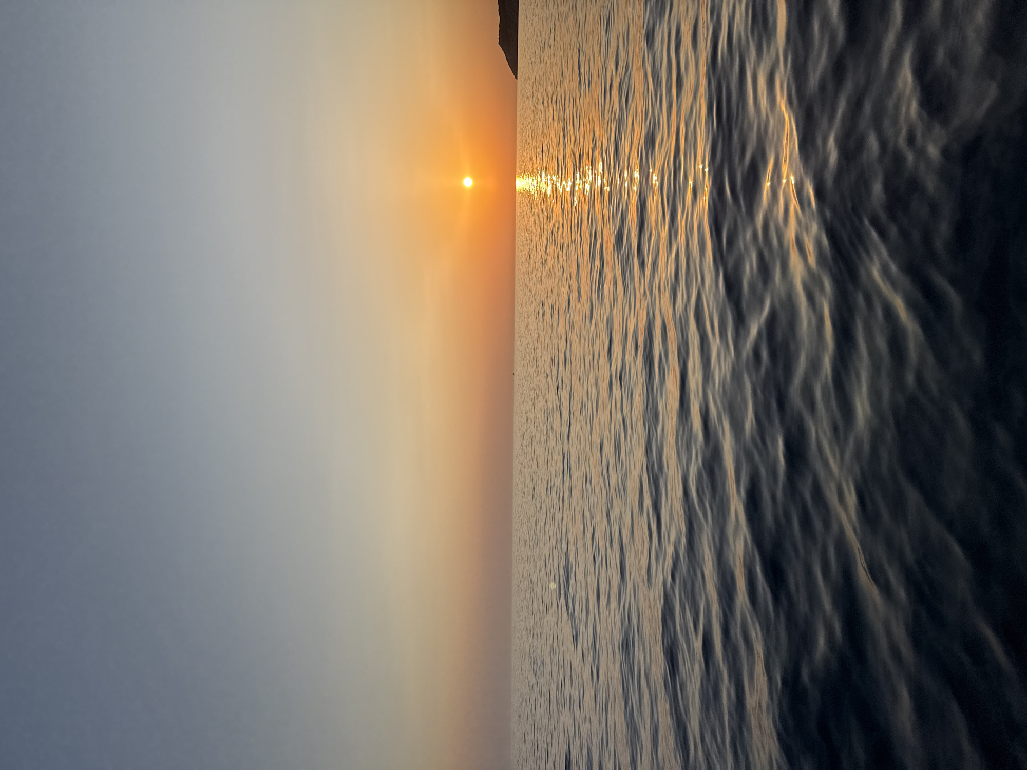 A serene sunset over the Adriatic Sea in Montenegro. The sky transitions from a pale blue at the top to a warm orange glow near the horizon, where the sun, a bright orb, is setting. The sun's reflection creates a shimmering path of light across the water. The sea is calm with gentle ripples, dark blue in the foreground, gradually lightening towards the horizon. In the far distance, a silhouetted island or coastline is visible on the right. There are no people or objects in the water, only the vast expanse of sea and sky.