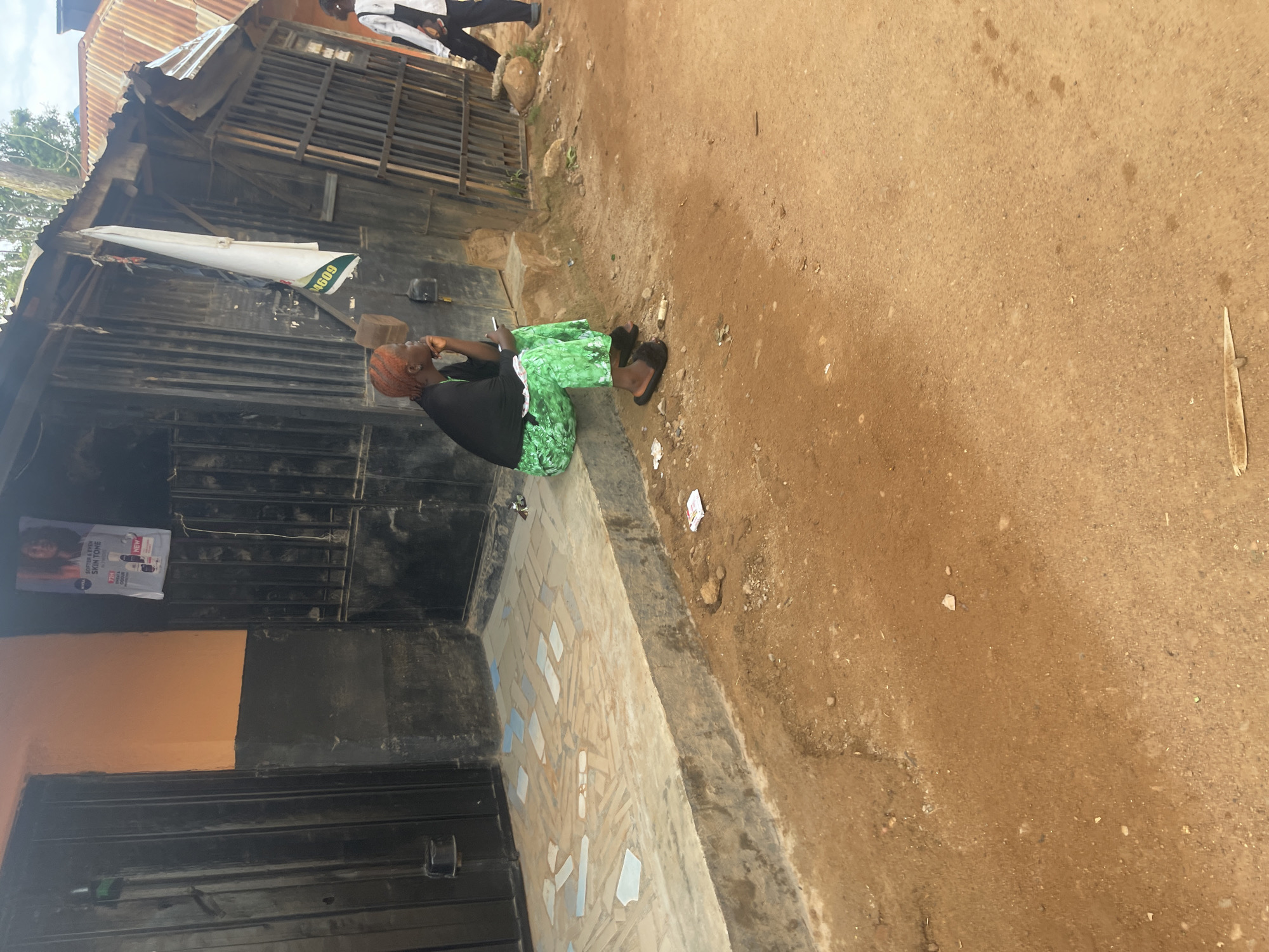 In Rafin Par, Nigeria, a woman sits on a concrete step in front of a weathered storefront. She wears a black top and vibrant green patterned trousers, with her hair dyed a striking orange. Her hand is raised to her chin, in a contemplative pose. She is wearing black fuzzy slippers. The storefront itself is constructed from dark metal, with bars on the windows and doors. Above the entrance, a faded poster with an image of a woman and product advertisements is affixed to the wall. A white cloth, possibly a flag, hangs from the corrugated metal roof. The ground in front of the building is made of reddish-brown dirt, with some scattered debris, including a small white packet. To the right, another person in a white top and dark trousers walks away from the scene. The overall impression is one of a typical urban scene in a developing country, with elements of daily life and commerce.