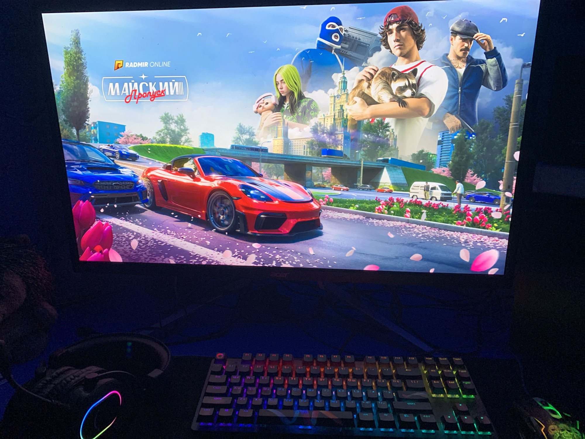 A monitor displays a digital image featuring stylized characters with animals superimposed over an urban landscape. A red Porsche and a blue Subaru are parked on a road with pink tulips and falling petals. Overhead are superimposed images of people, including two men holding raccoons, a woman with green hair, and a figure with a blue ski mask holding a boombox. In the background is a cityscape with a bridge. The monitor sits on a desk, where a gaming keyboard with RGB lighting is visible, as well as headphones and a gaming mouse. The location is not identifiable as Severodvinsk, Russia, based on the image content.