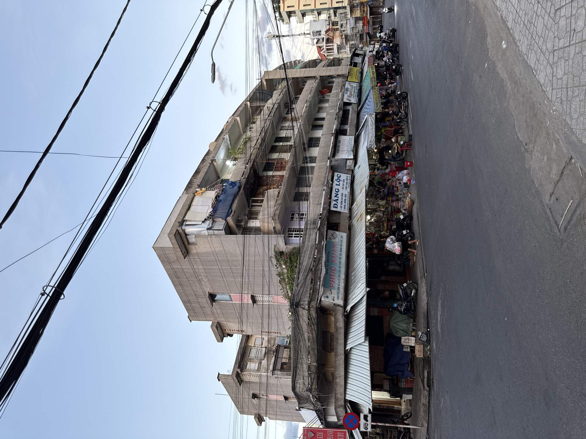 This is a street-level, medium-distance, eye-level shot of a busy urban intersection in Nha Trang, Vietnam, during the day, possibly late afternoon or early evening, given the soft light. The primary subject is a multi-story, utilitarian apartment building on the left, with a row of market stalls and shops operating on the ground floor and a lower, attached structure that extends out over the sidewalk.
The building itself appears aged, constructed with concrete and exhibiting a somewhat brutalist architectural style. It has multiple balconies and windows, some of which are adorned with laundry or personal items, indicating residential occupancy. The building is heavily draped with a dense network of overhead electrical wires, typical of Vietnamese cities.
Below the apartment building, a covered walkway shelters various shops and stalls. These are stocked with an array of goods, including what appear to be household items, baskets, and other merchandise, suggesting a local market or shopping area. Awnings, both striped and plain, provide shade.
Several motorcycles are parked along the sidewalk and street, and a few individuals are visible. On the left, a person is walking away from the camera, carrying a white plastic bag. To the right, more people are browsing or tending to the shops. A motorcycle is also seen being ridden down the street on the far right.
The street itself is paved and appears to be a main thoroughfare. A traffic sign with red and blue circles and a white symbol is visible on a pole on the left side of the image, indicating traffic regulations. The overall environment is one of urban density and everyday commerce. The sky above is a pale blue with some wispy clouds.