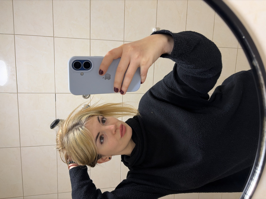 An image captures a mirror selfie of a young woman, rotated 90 degrees clockwise. The woman has light blonde hair, parted slightly off-center, with some strands falling across her forehead and face. Her eyes are open and looking directly at the camera, and her lips are softly parted, showing a natural, light pink hue. She wears a dark, possibly black, long-sleeved top or sweater with a textured, slightly fuzzy fabric and a high neckline. A dark earring is visible in her left ear, and a dark bracelet or band can be seen on her left wrist.
Her left hand holds a light blue or grey smartphone with a case, and the Apple logo is visible on the back of the phone. Two camera lenses are prominent on the top left of the phone's back. Her fingernails are painted with a dark red or burgundy polish. Her right arm is bent, with her hand or forearm resting near her head or hair.
The setting reflected in the mirror appears to be an indoor space with light-colored, possibly beige or cream-colored, square tiles on the wall behind her. A small, dark cylindrical object, possibly a hook or part of a fixture, is visible on the tiled wall in the reflection. The mirror itself is round, with a distinct black frame visible on the right side of the image. The lighting is even, suggesting an indoor setting without direct sunlight.
