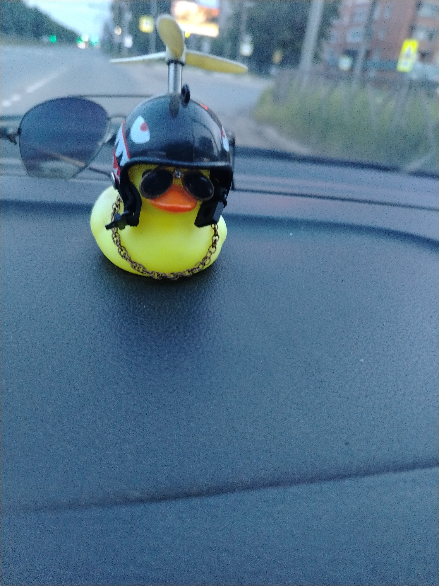 A yellow rubber duck wearing sunglasses and a helmet with a propeller on top sits on the dashboard of a car. A pair of sunglasses are placed adjacent to the duck, also resting on the dashboard. The backdrop suggests an outdoor scene in an urban environment, potentially in Fedorino, Russia. A road with greenery and buildings alongside is visible in the distance.
