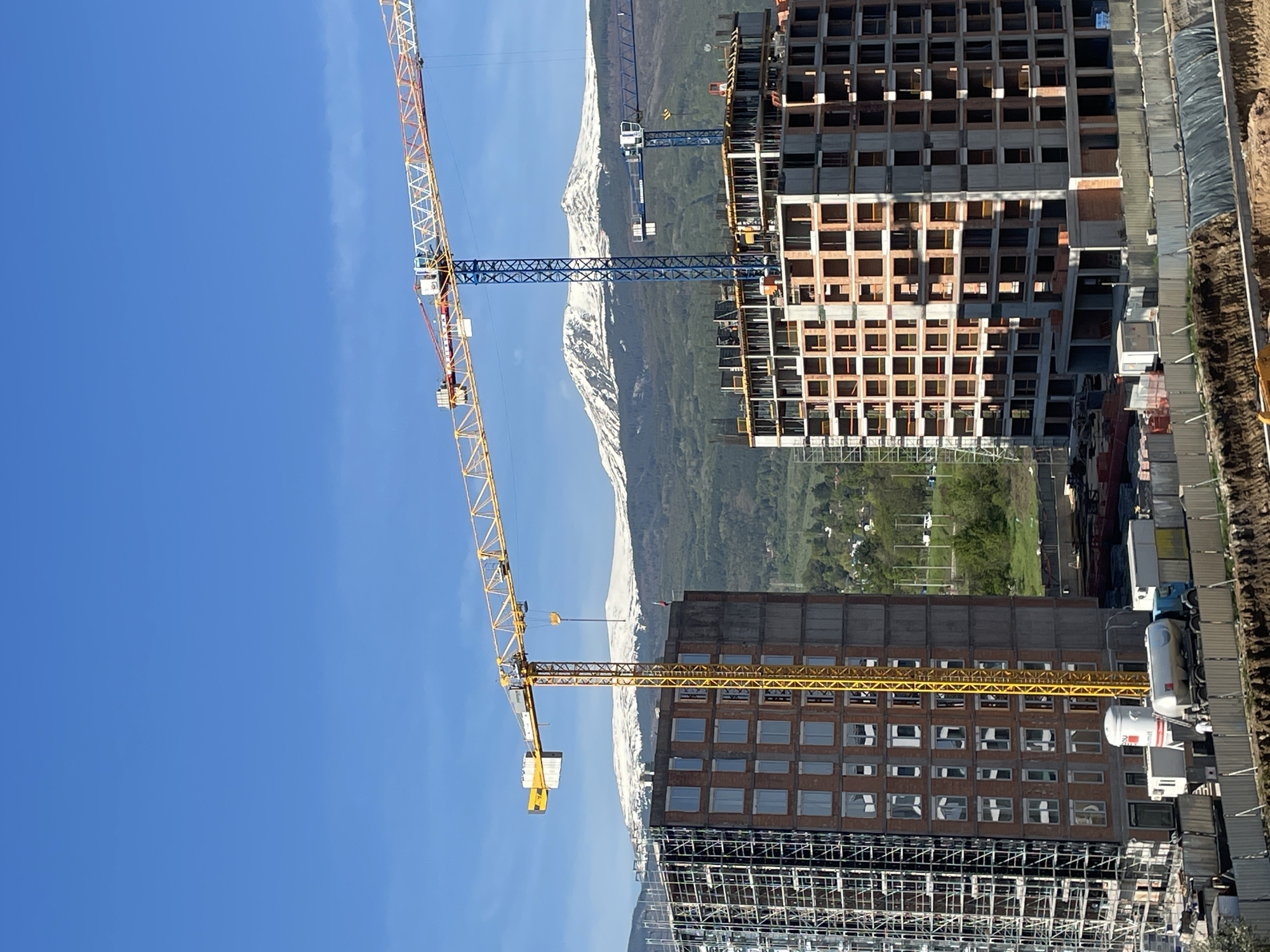 The image shows a construction site in Sofia, Bulgaria, with two buildings under construction. Two tower cranes, one yellow and one blue, are visible. The backdrop includes snow-capped mountains and green trees. A clear blue sky is seen above. Scaffolding surrounds the lower part of one of the buildings. Construction materials are visible near the base of the buildings.
