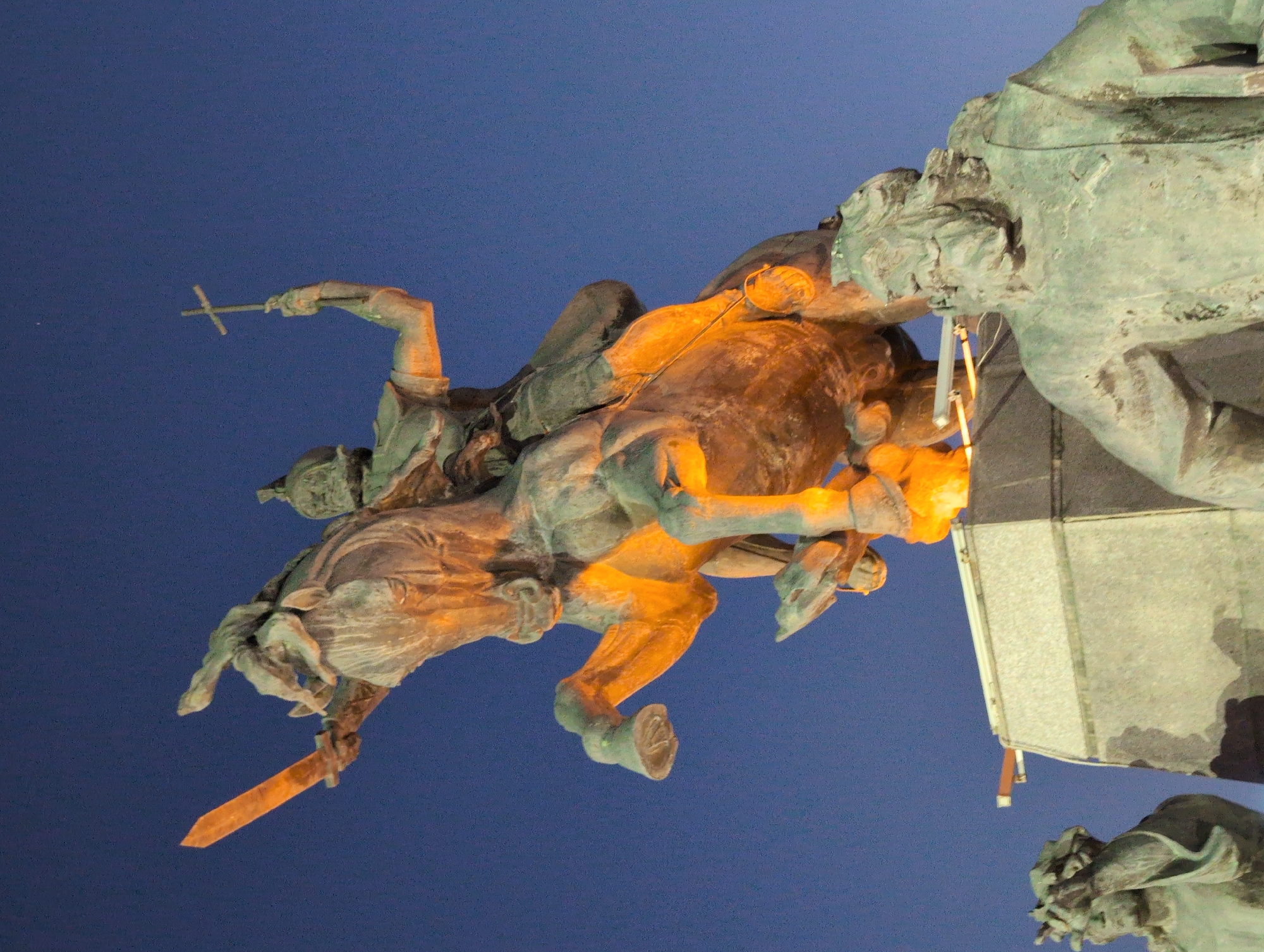 This is a nighttime photograph of the statue of Vardan Mamikonian in Gyumri, Armenia. The statue depicts Vardan Mamikonian, a military leader and saint in the Armenian Apostolic Church, on horseback. He is shown raising a sword in his right hand and a cross in his left hand. The equestrian statue is made of bronze and is illuminated by artificial lights, giving it a golden hue against the dark blue sky. In the foreground, parts of other statues are visible, appearing to be on a lower level or at the base of the monument. The statue is located in a plaza or public space, with architectural elements of the pedestal and surrounding structures visible. The sky is clear and devoid of clouds, suggesting a calm night.