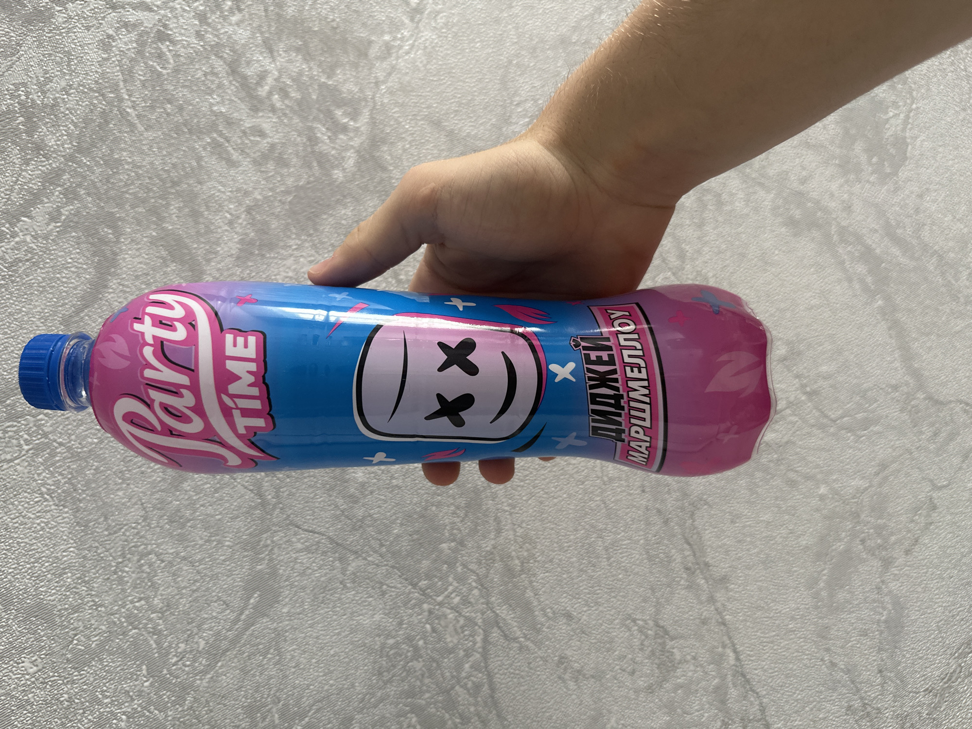 A hand holds a bottle of "Party Time" drink. The bottle is blue with pink and purple accents, and features the image of a marshmallow with X's for eyes and a smile. Text on the bottle reads "Party Time" and "DJ Marshmallow". The background is a textured, light-colored wall. The location is the city of Birobidzhan, Russia.