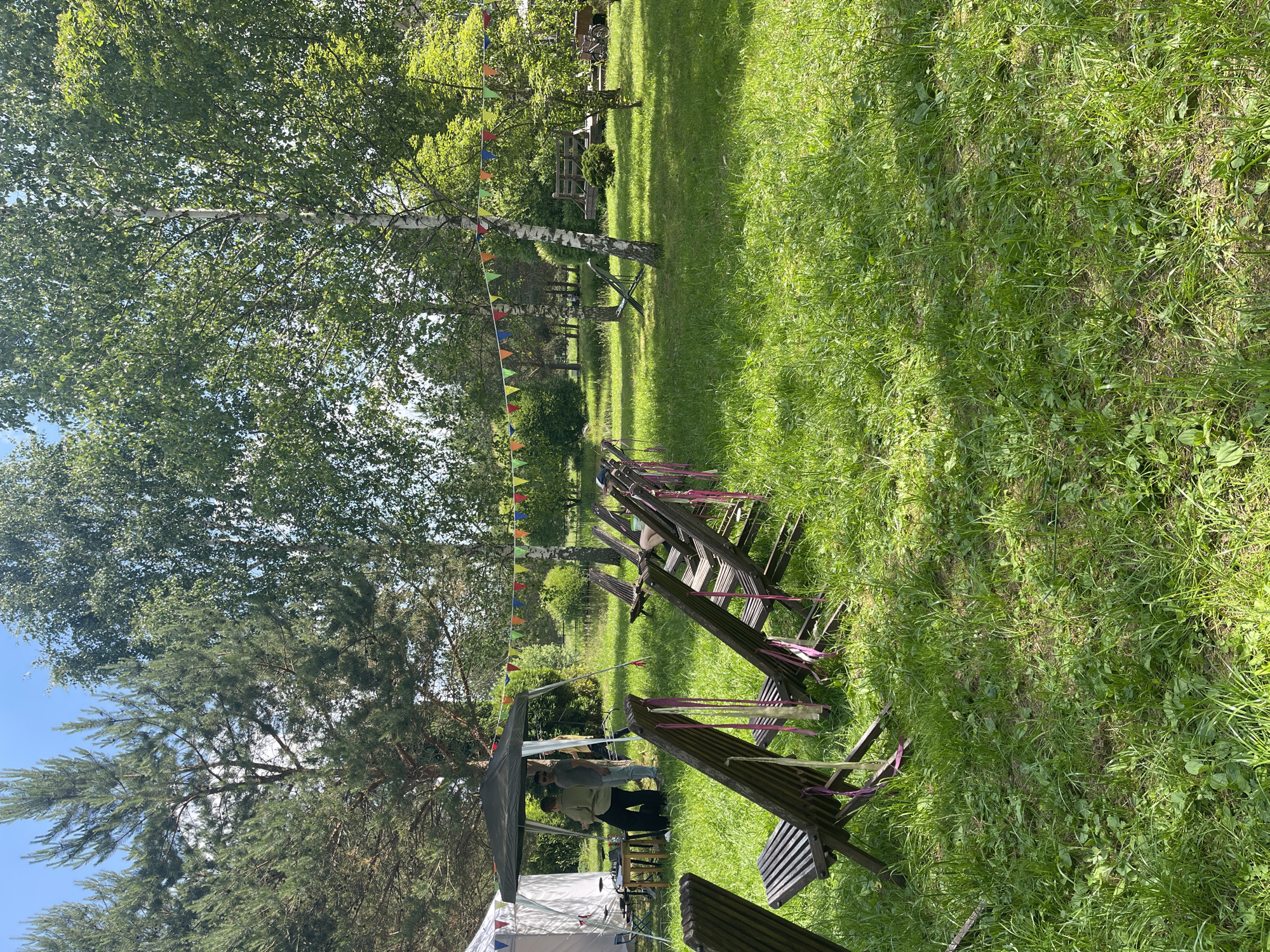 An outdoor scene depicts a grassy field with several wooden chairs arranged in rows. A string of colorful triangular flags stretches across the frame between trees. In the background, a black canopy tent is set up, with two individuals standing beneath it. To the left, a white tent is also visible. The overall environment appears to be a park or outdoor event space, characterized by lush green grass and numerous trees.