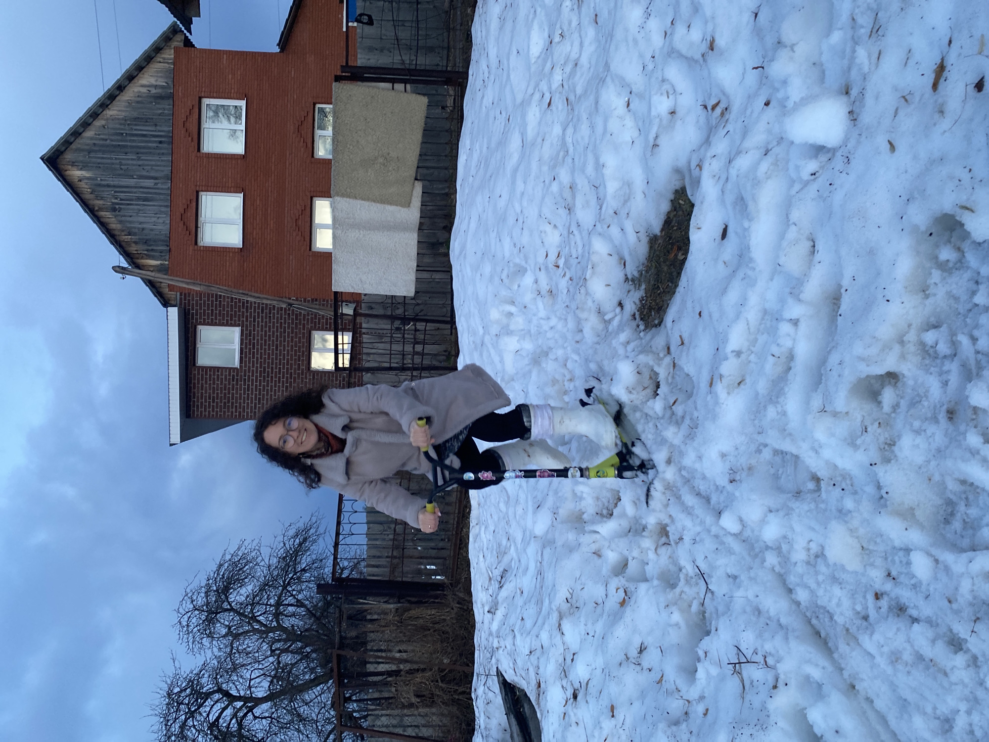 The image shows a woman riding a scooter in a snowy area near a brick building in Ольховка, Russia. The woman has dark curly hair and wears glasses, a light-colored coat, and dark leggings. She is on a scooter with a black body and a green-yellow steering mechanism. The ground is covered in snow with some patches of dry ground visible. A metal fence runs along the edge of the snow, and a bare tree is seen to the left of the fence. A two-story brick building with a wooden gable roof and several windows is in the background. Two rugs hang on a clothesline attached to the building. The sky is overcast.