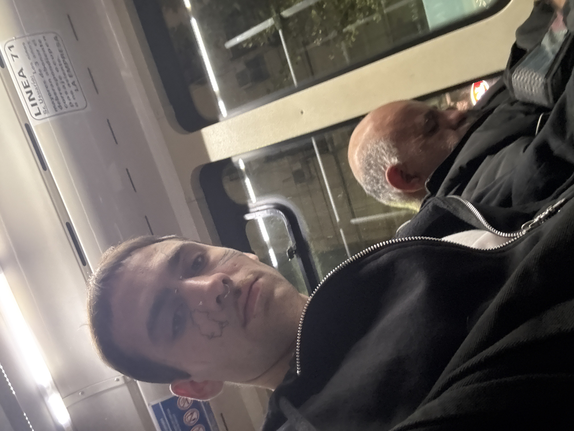 The image depicts a selfie taken inside public transport. The subject, a young man, is visible in the foreground, looking directly at the camera. There is a visible tattoo near his left eye. In the background, an older man is asleep. The sign "LINEA 71", indicating the bus line number, can be seen inside the vehicle, indicating location in Buenos Aires, Argentina. The reflection in the bus window shows some buildings and trees outside. Both individuals are dressed in dark clothing.