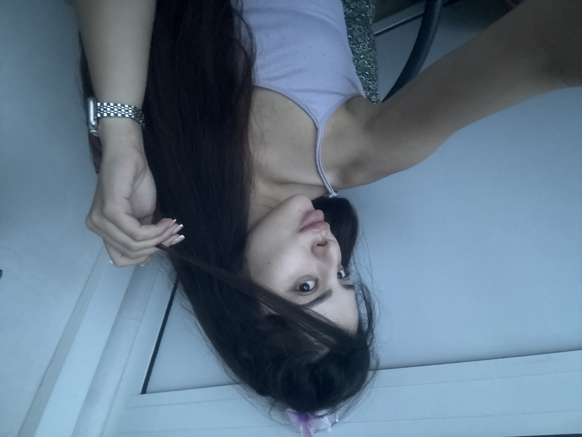 The image is an indoor portrait of a young woman, rotated upside down, featuring her upper body. She has long, dark brown hair that frames her face and flows downwards in the rotated orientation. Her gaze is directed towards the camera with a neutral expression, characterized by slightly parted lips and wide, dark eyes.
She is wearing a light purple, ribbed tank top with thin shoulder straps. On her left wrist, she wears a silver-toned smartwatch with a patterned, checkered metallic band. Her left hand, with fingernails tipped in white, is raised near her head, seemingly touching or playing with a strand of her hair. A small, faint light purple or pink ribbon or hair accessory is visible amidst her hair towards the bottom of the frame.
The background consists primarily of a plain, light blue or grey wall or ceiling. White architectural elements, possibly window frames or wall paneling, are visible along the left and bottom edges of the main image. A small portion of a green fabric with a subtle floral pattern can be seen behind her right shoulder. The lighting is soft and ambient, suggesting an indoor setting without indicating a specific time of day. No discernible text is visible in the image.