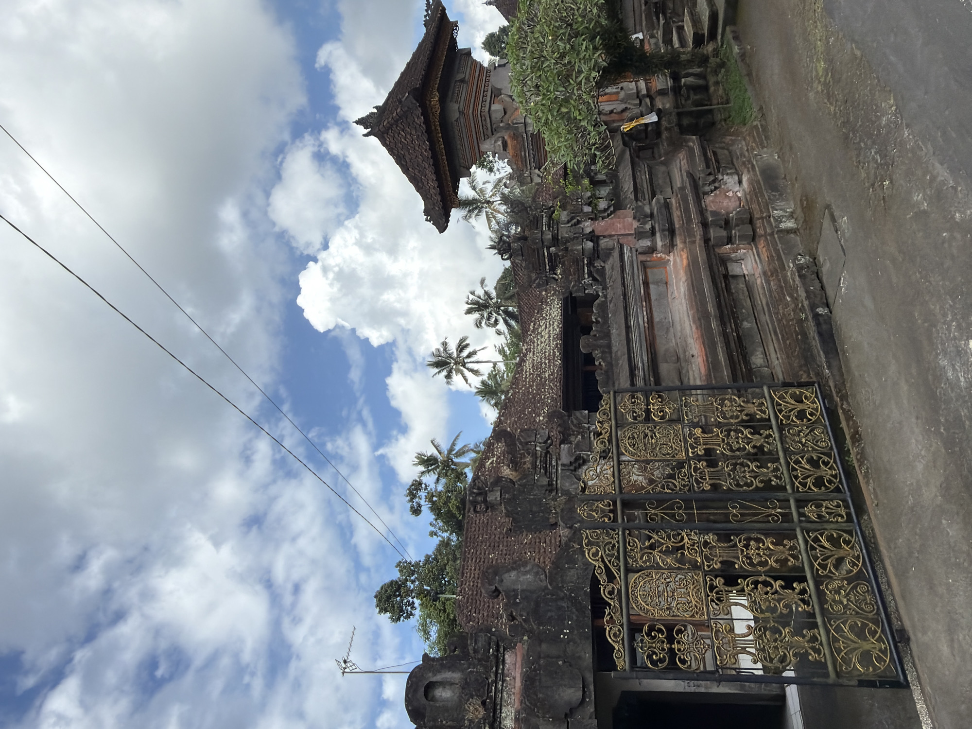 The image shows an ornate, dark stone gateway with intricate gold detailing, typical of Balinese architecture. The gate is partially open, revealing a glimpse of what might be an interior courtyard or building beyond. The gateway itself is ancient, with weathered stone and moss growing in crevices, suggesting age and a tropical climate. Above and behind the gate, a traditional Balinese temple structure is visible, characterized by its tiered, dark tiled roof and intricately carved facade. Lush green trees and palm fronds peek over the temple walls, indicating a tropical environment. The sky is a vibrant blue, dotted with fluffy white clouds, suggesting it is daytime with fair weather. Two dark power lines stretch diagonally across the sky, a modern element contrasting with the ancient structures. A small, modern antenna is visible on a pole in the lower left quadrant of the image. The scene evokes a sense of cultural heritage and tranquility, with the architecture and natural elements blending together. The location is identified as Tampaksiring, Indonesia, known for its ancient temples and archaeological sites.