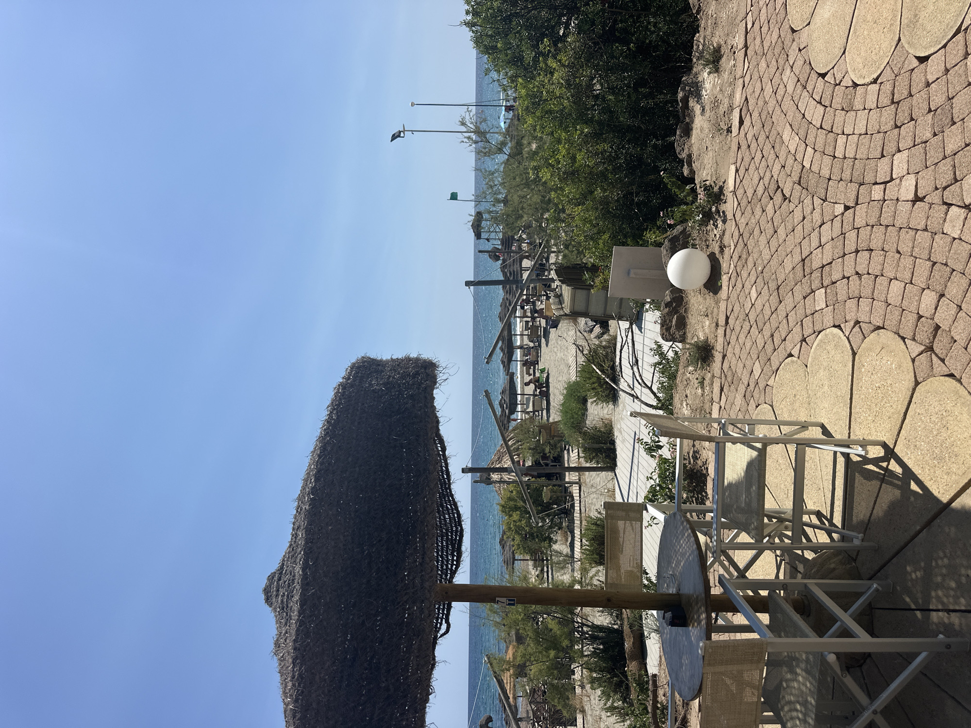 This image captures a serene beachside scene in Biddeputzi/Villaputzu, Italy, under a clear blue sky. In the foreground, a thatched umbrella shades a round wooden table accompanied by two white director-style chairs. The table has a small ashtray on it. The seating area is situated on a paved patio made of tan, irregularly shaped stones, bordered by a cobblestone path.
Beyond the immediate seating area, the sandy beach stretches out towards the vibrant blue sea. Numerous beach umbrellas and loungers are visible, suggesting a public beach with amenities. Lush green bushes and trees are scattered along the shoreline and further inland, adding a touch of nature to the scene. A few people can be seen relaxing on the beach in the distance.
The overall impression is one of a tranquil summer day at the beach, ideal for relaxation and enjoying the coastal Italian atmosphere.