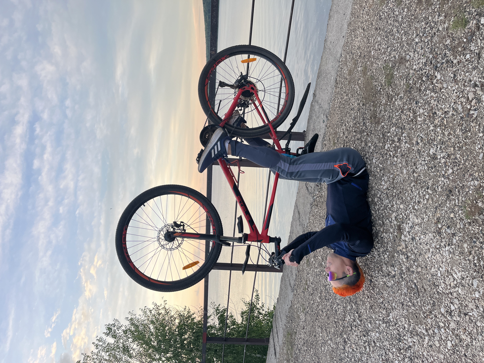 A man with dyed orange hair lies on a gravel surface next to a red bicycle leaning on a metal railing. The man wears sunglasses, a blue long-sleeved shirt, and grey pants with a stripe down the side. His feet are in athletic shoes. The bicycle is red with black accents and has two visible wheels. The railing separates the gravel area from a body of water. Beyond the water, the sky is visible with clouds. The location is Chaikovsky, Russia.
