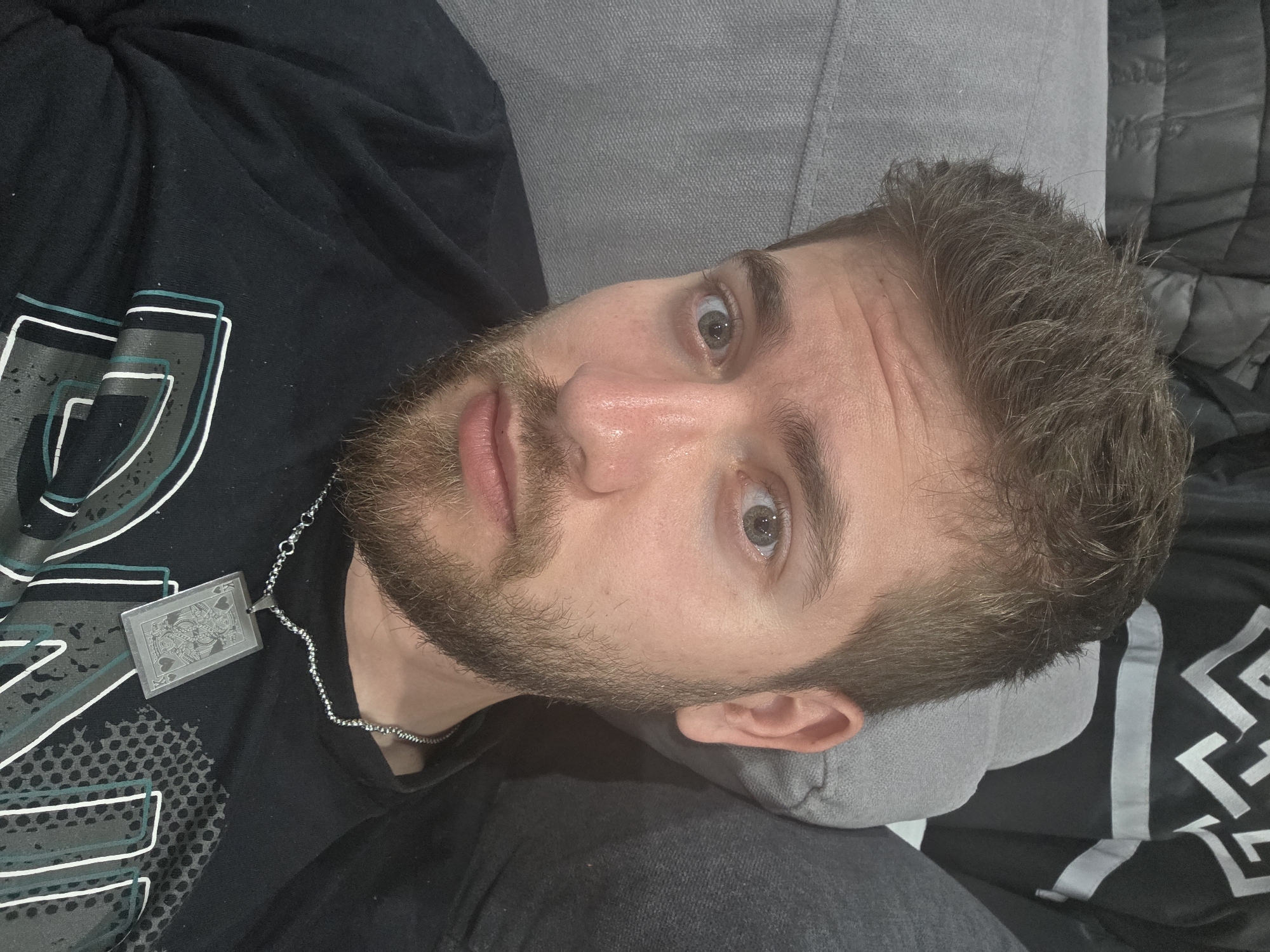 A close-up, angled shot captures a man with fair skin and a beard, possibly in his late 20s to early 30s. He is lying down, with his head resting on a light gray pillow. His hair is short, textured, and a medium brown color. He is wearing a black t-shirt with a graphic design featuring geometric shapes and possibly text in shades of white, gray, and teal. A silver chain necklace with a playing card pendant (the Queen of Spades) is visible around his neck. His eyes are open and appear to be light gray or blue. The background includes what appears to be a dark gray upholstered sofa or bed. There is no information to suggest the image was taken in Yagodovo, Bulgaria.
