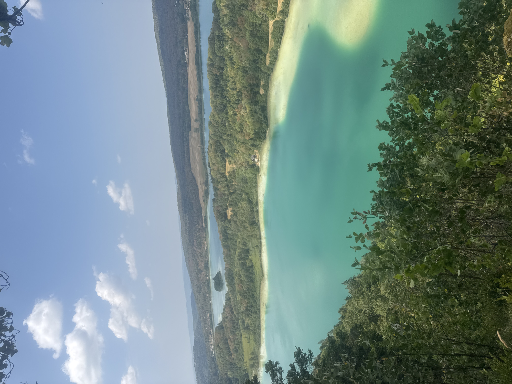 The image captures a panoramic view of a vibrant turquoise lake set against a backdrop of rolling, tree-covered hills and a clear blue sky with scattered white clouds. In the foreground, lush green foliage partially obscures the view, adding depth to the scene. The lake's surface exhibits a gradient of colors, transitioning from a deep turquoise in the foreground to a lighter, almost milky hue near the shoreline, suggesting shallows or submerged sediment. Further in the distance, another body of water is visible, with a small, tree-covered island nestled within it. The landscape appears serene and natural, indicative of a rural or park-like setting. The location is identified as the Lac de Chalain, situated in the commune of La Chaux-du-Dombief, France.