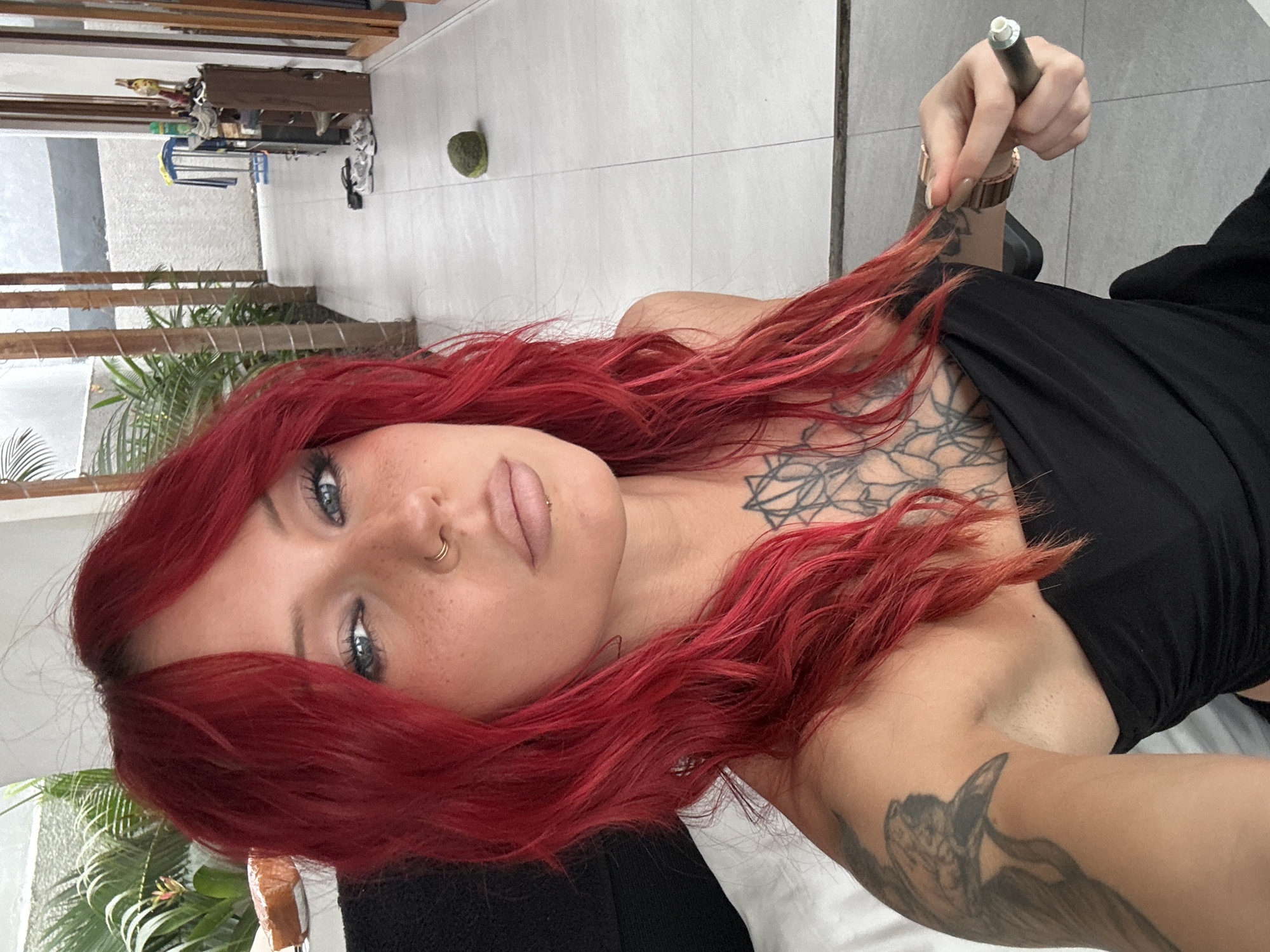 A woman with vibrant red hair, fair skin, and blue eyes is captured in a selfie taken in what appears to be an outdoor hallway of a residential property in North Kuta, Indonesia. She is wearing a black strapless top and has tattoos on her chest and left arm. Her right hand is holding a vape pen. The background shows a tiled floor, a walkway with wooden beams, lush green plants, and a glimpse of furniture and personal belongings on shelves. The lighting suggests it is daytime, possibly with natural sunlight filtering into the area.