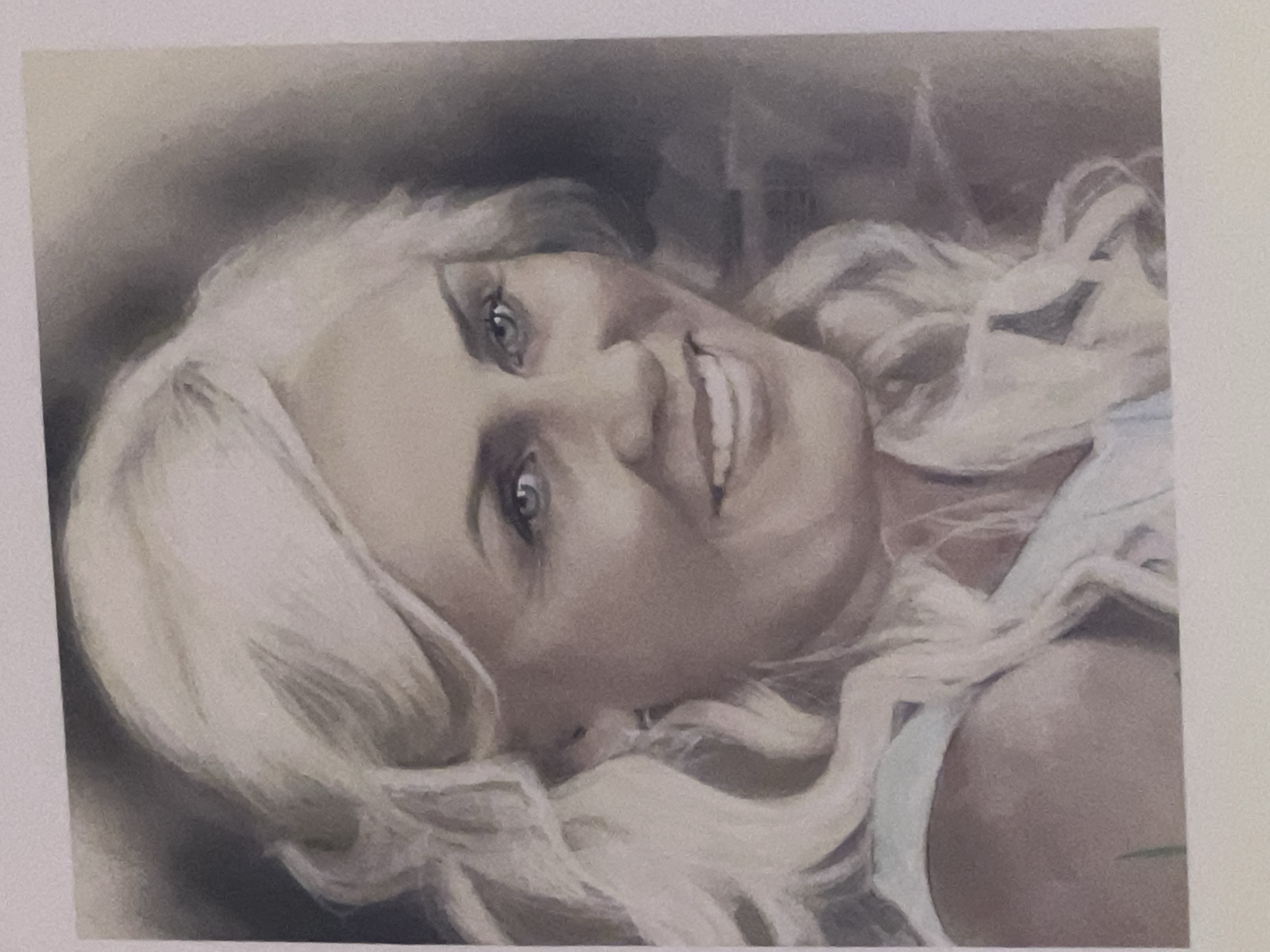 The image is a portrait of a blonde woman with blue eyes. She has long, wavy blonde hair and is smiling. She is wearing a light blue top. The background is dark and out of focus. There is no information available about the location of Bobrovka, Russia in relation to this image.